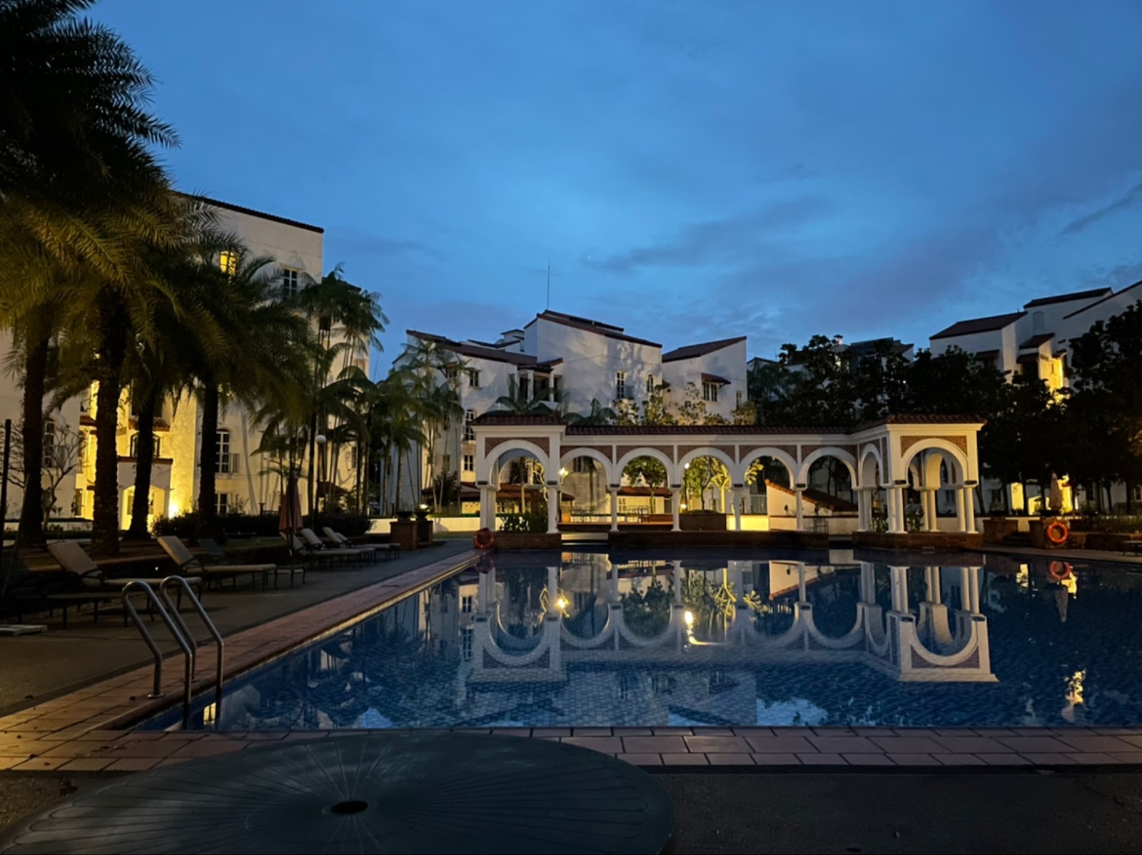 Andalucia: Twilight view of a resort-style swimming pool with an arched white pergola, palm trees, and surrounding residential buildings