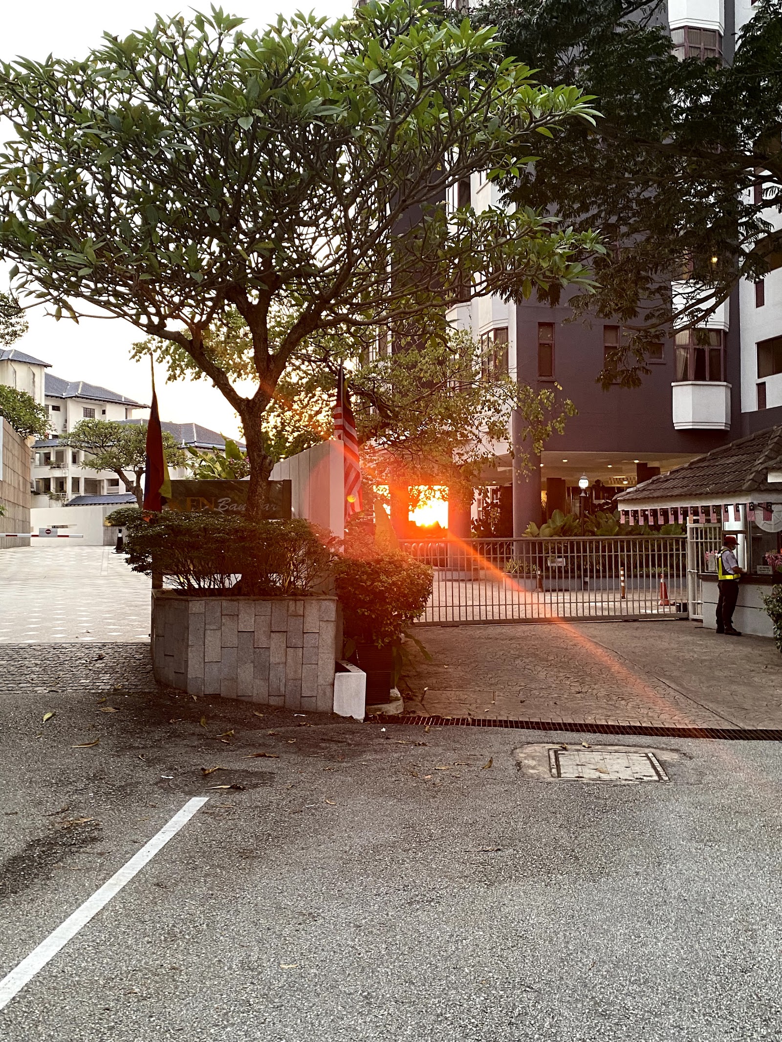 Desa Bangsar Ria: Entrance driveway and gate of a residential development at sunset, with a guardhouse, security guard, trees, and the building facade visible in the background