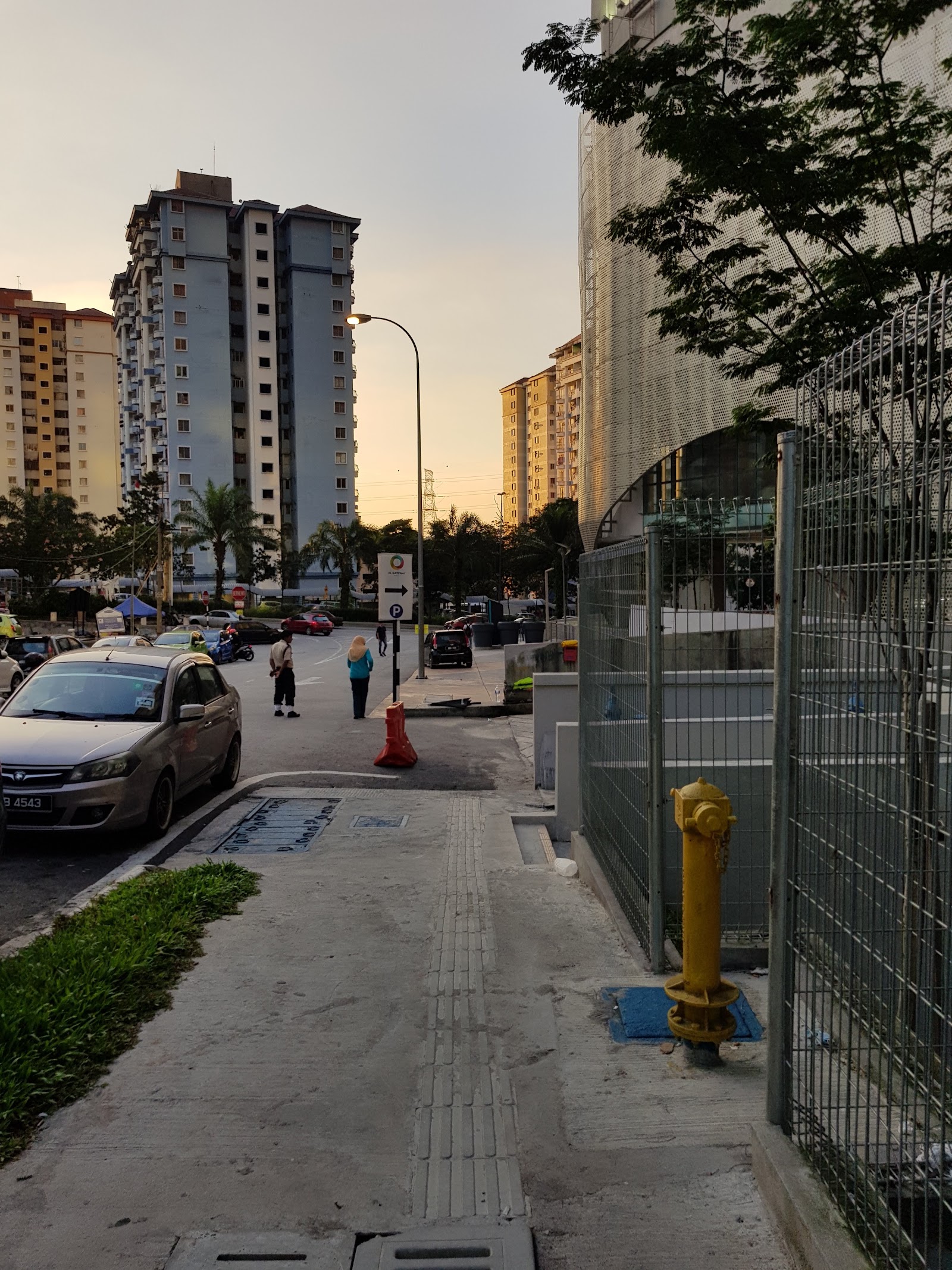 Residensi Kerinchi: Street view outside a residential development showing parked cars, pedestrians, high-rise towers, a fire hydrant and fencing along the sidewalk