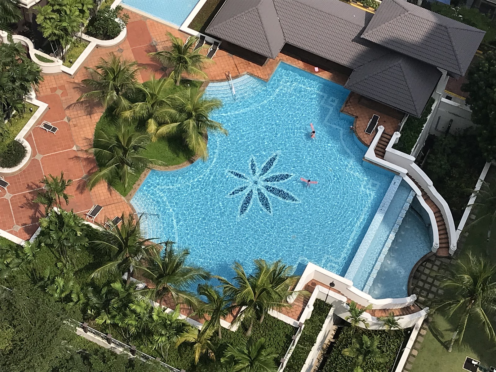Almaspuri: Aerial view of a resort-style swimming pool with a decorative flower mosaic, surrounded by palm trees, sun loungers and a tiled courtyard, with two swimmers in the water