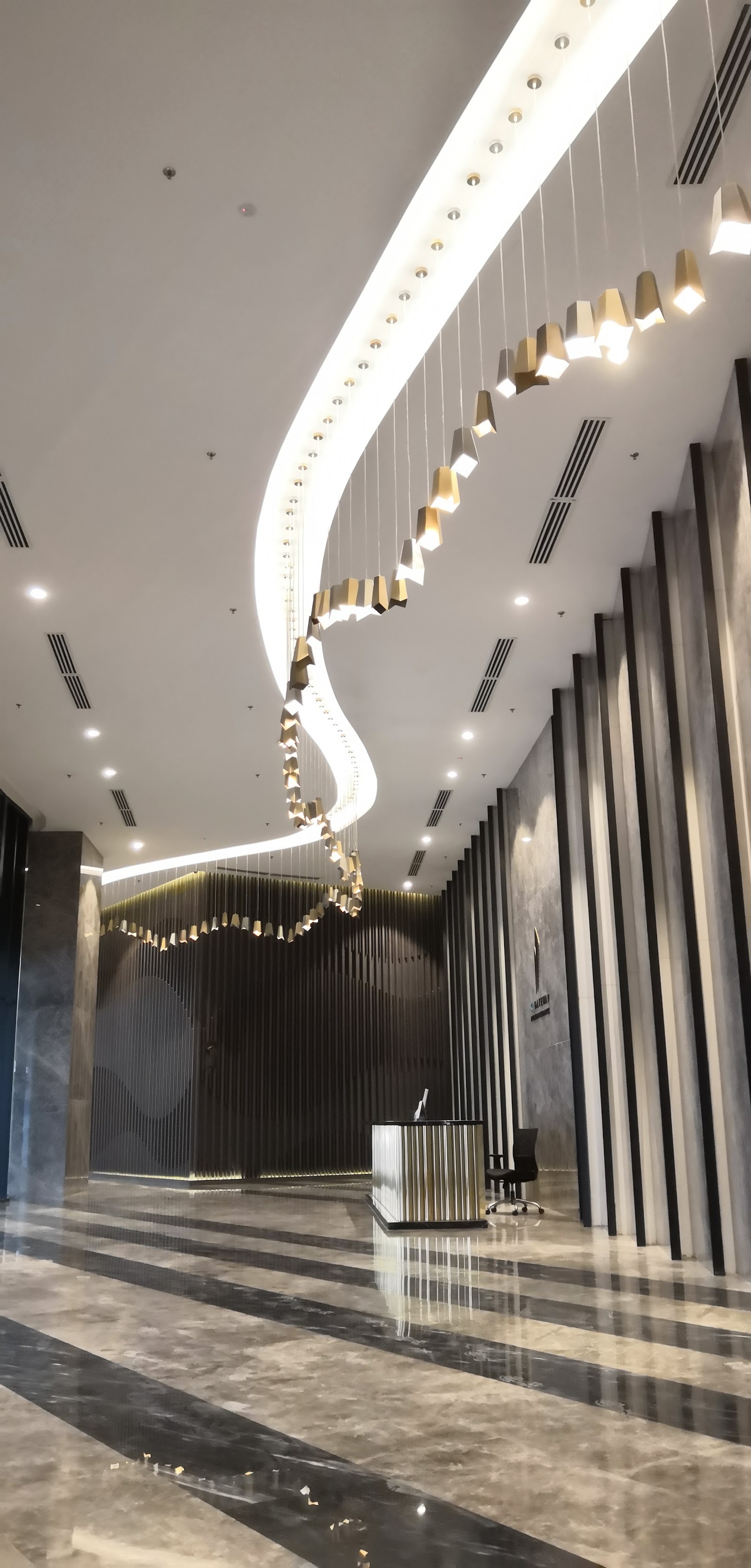 KL Gateway, Premium Residences: Elegant hotel-style lobby with a sculptural spiral chandelier hanging from the ceiling, dark vertical panel wall, marble flooring, and a reception counter