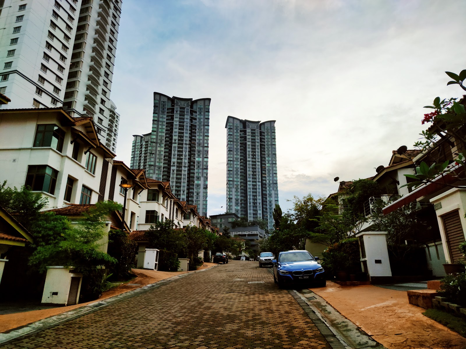 Kiaramas Sutera: A residential street lined with townhouses leading toward tall high-rise condominium towers, with a car parked on the paved road.