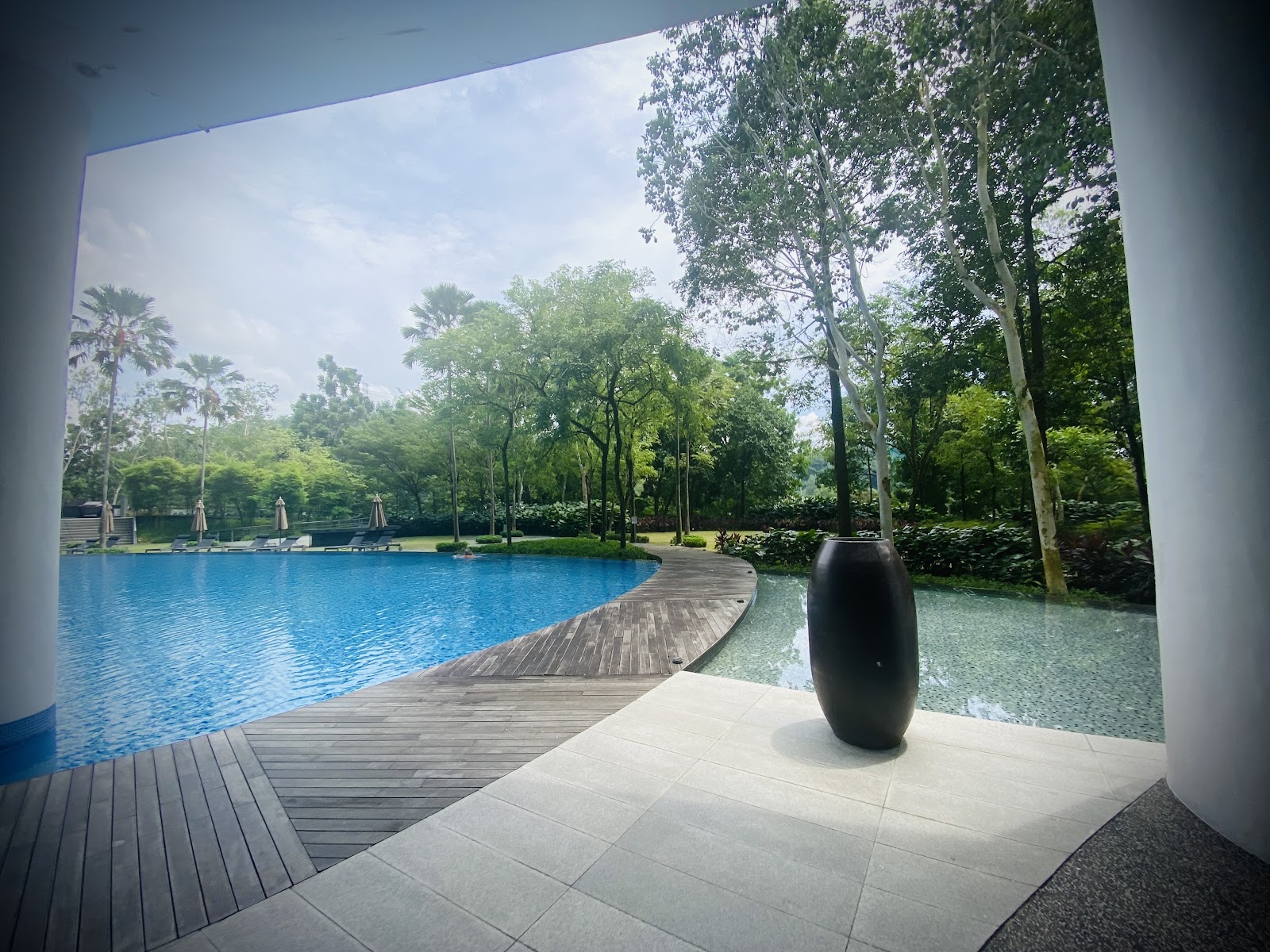 Sunway Palazzio: Infinity-edge swimming pool with wooden deck surrounded by lush greenery, viewed from a covered walkway with a large black decorative vase