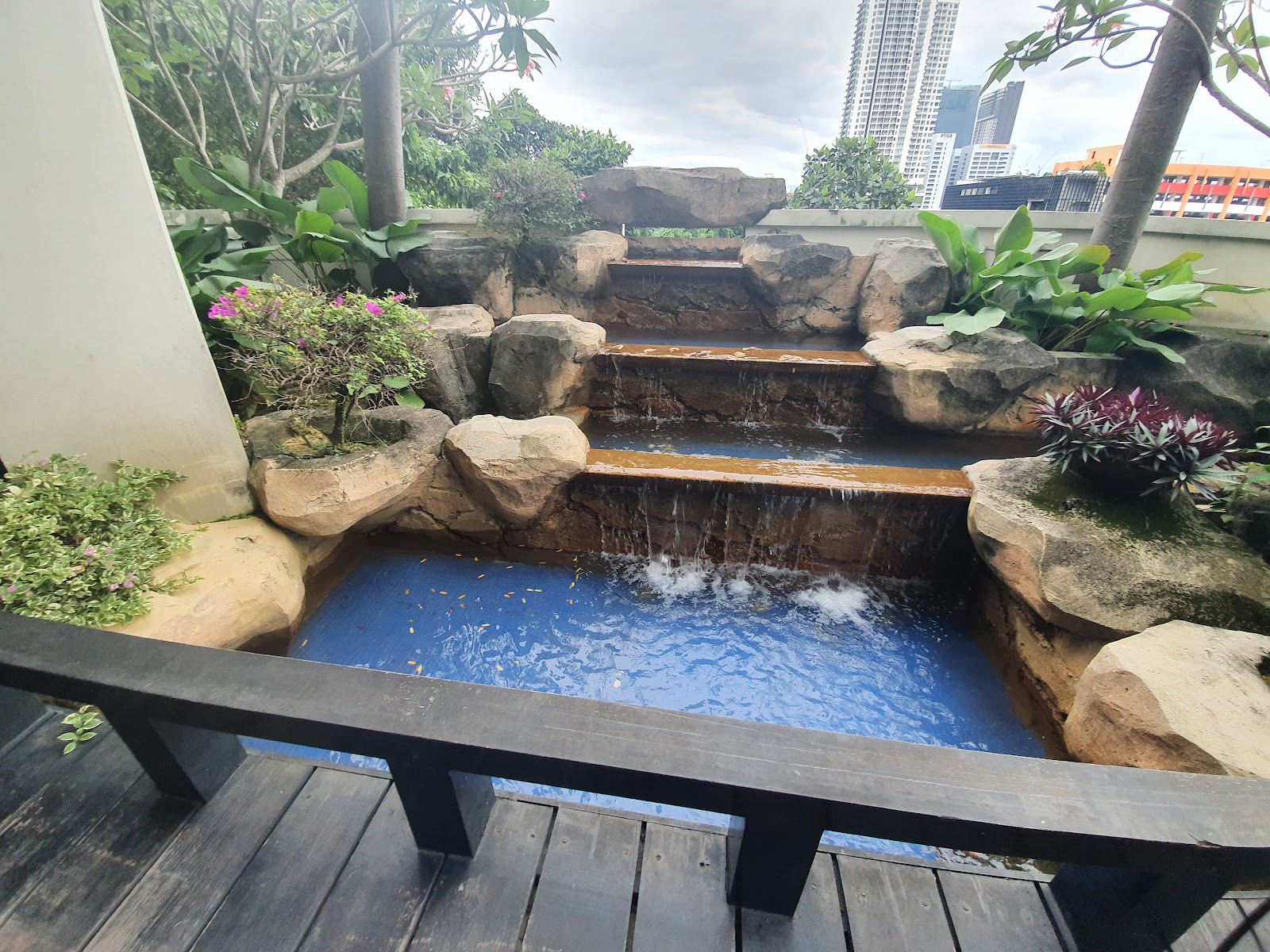 Suasana Bangsar: A landscaped waterfall feature with rock formations and a wooden deck, part of a residential development's outdoor amenities, with city skyline in the background