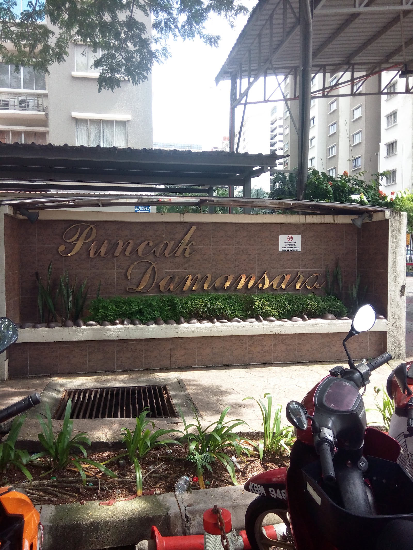 Puncak Damansara: Entrance signage wall for Puncak Damansara with gold lettering, landscaping, motorbikes parked in front, and residential towers in the background