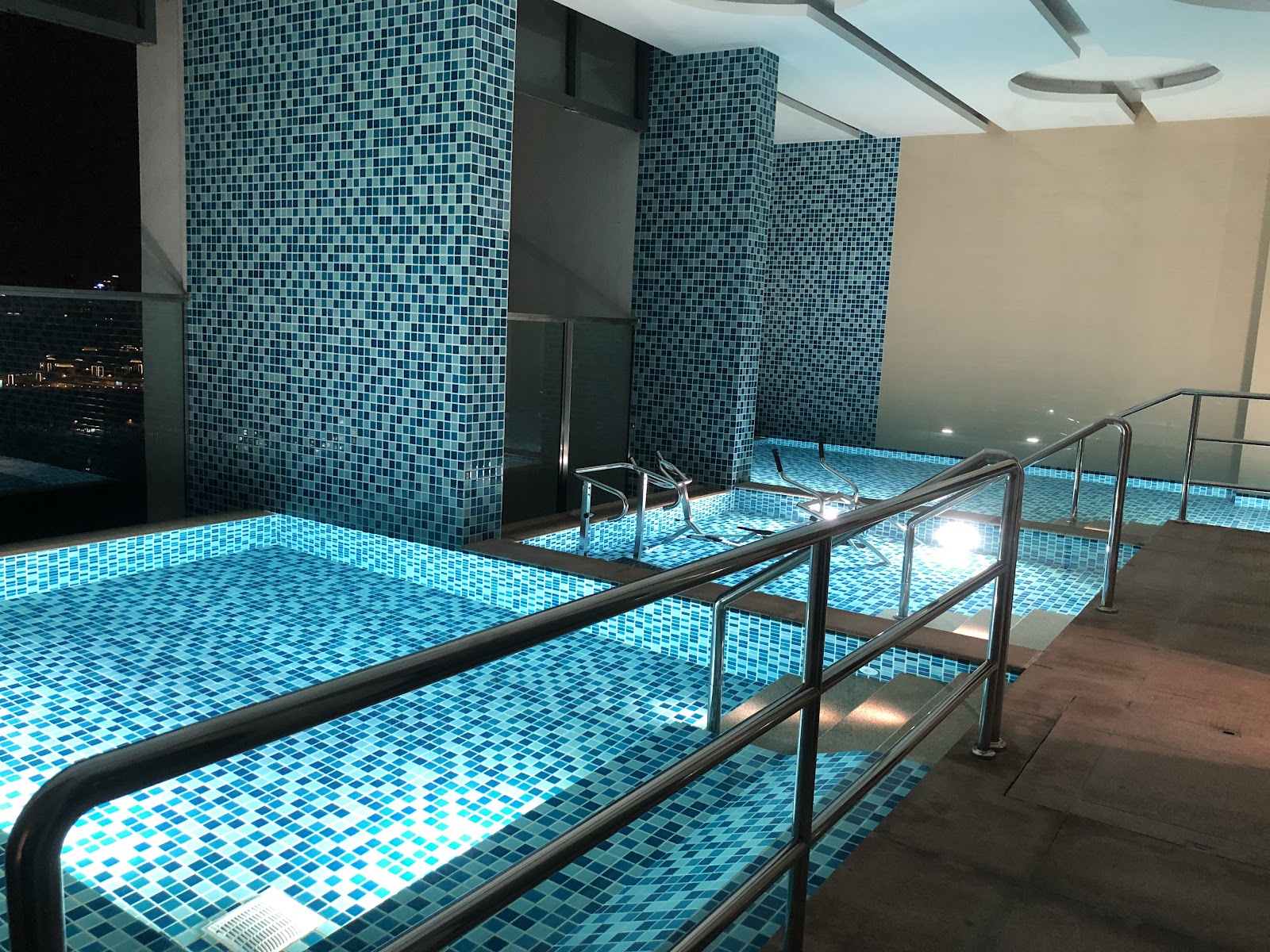 Agile Mont Kiara: Indoor infinity-style pool and jacuzzi area with blue mosaic tiles, chrome handrails, and a night city view through the window