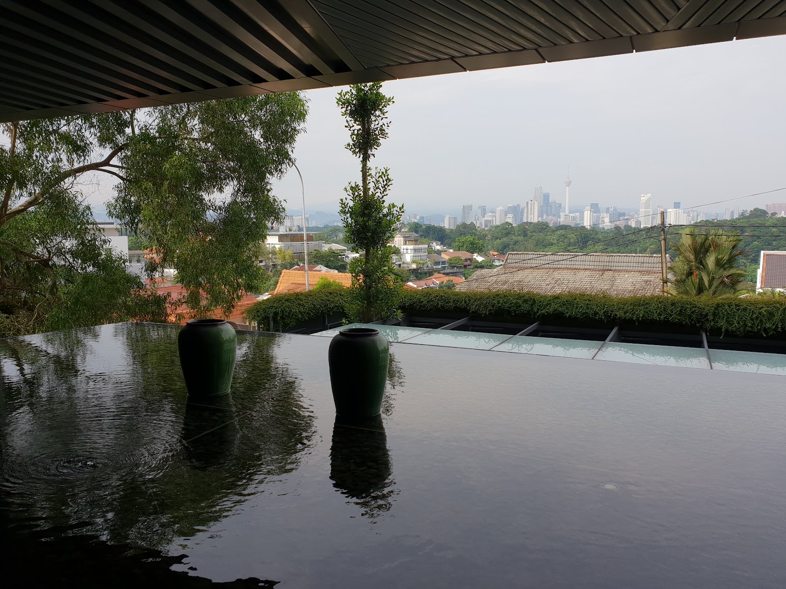 Rhombus @ Bangsar: Infinity pool edge with two dark ceramic vases overlooking a cityscape with skyline towers, rooftops, and trees