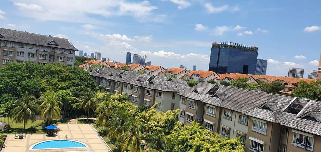 Sri TTDI: Aerial view of a low-rise townhouse development with tiled roofs, surrounded by trees, a swimming pool, and city skyline in the background