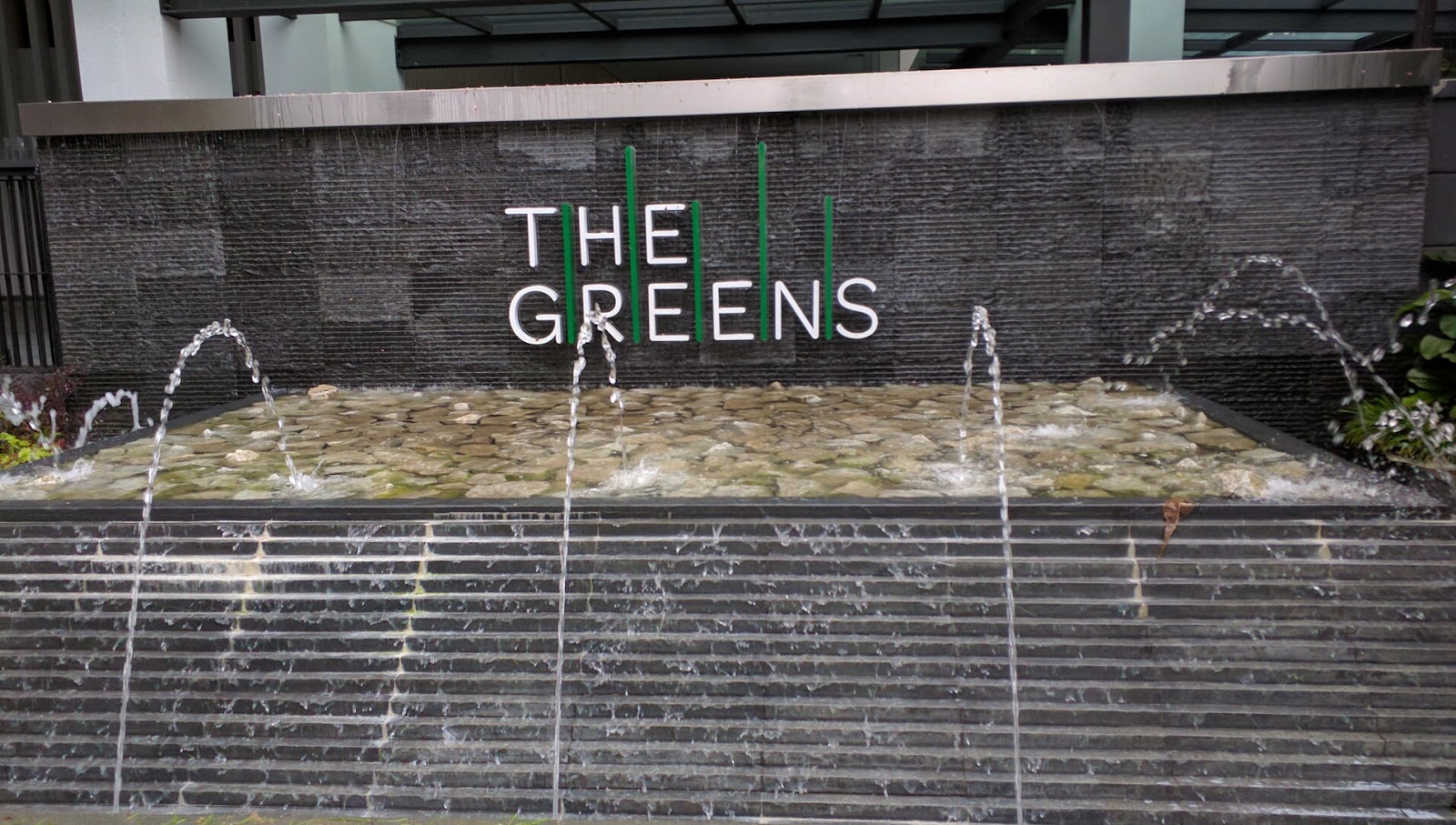 The Greens: Fountain feature with stepped water cascade in front of a stone wall bearing 'THE GREENS' signage