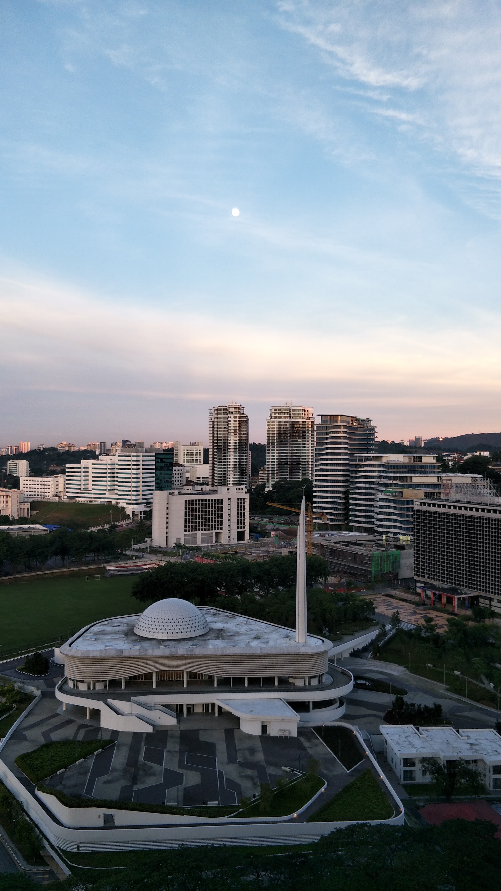 Pantai Towers: Aerial view of a city skyline with high-rise towers, a domed building with a spire in the foreground, and green fields, at dusk with a visible moon