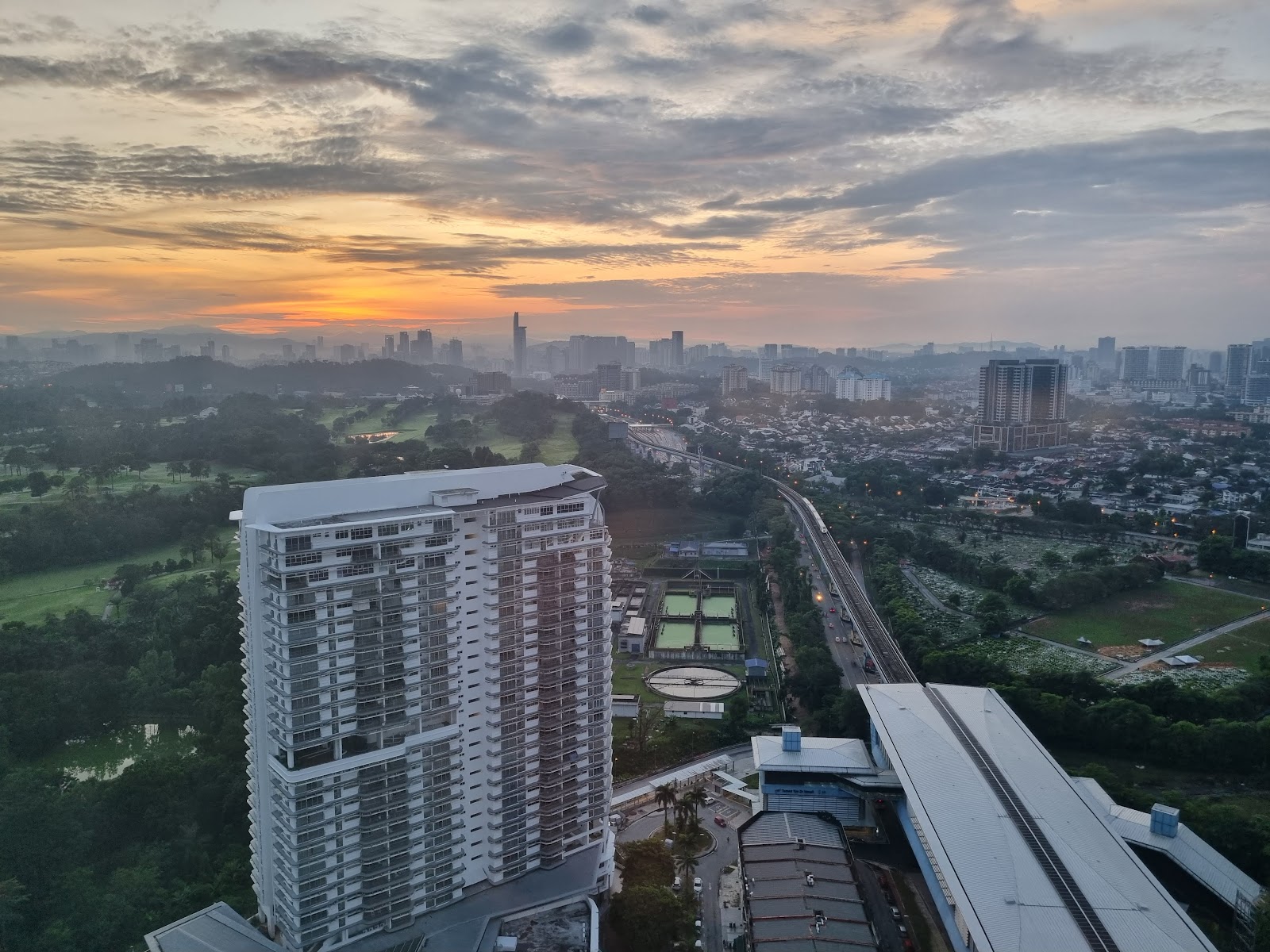 The Plaza Condominium: Aerial view of a high-rise residential tower at sunset with the city skyline, tennis courts, and a light rail track in the background