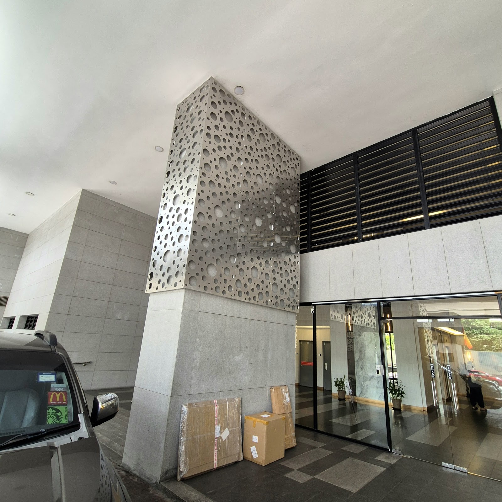 Glomac Centro: Entrance area of a building showing a decorative perforated metal panel feature, glass doors leading into a lobby, and boxes stacked near a parked car
