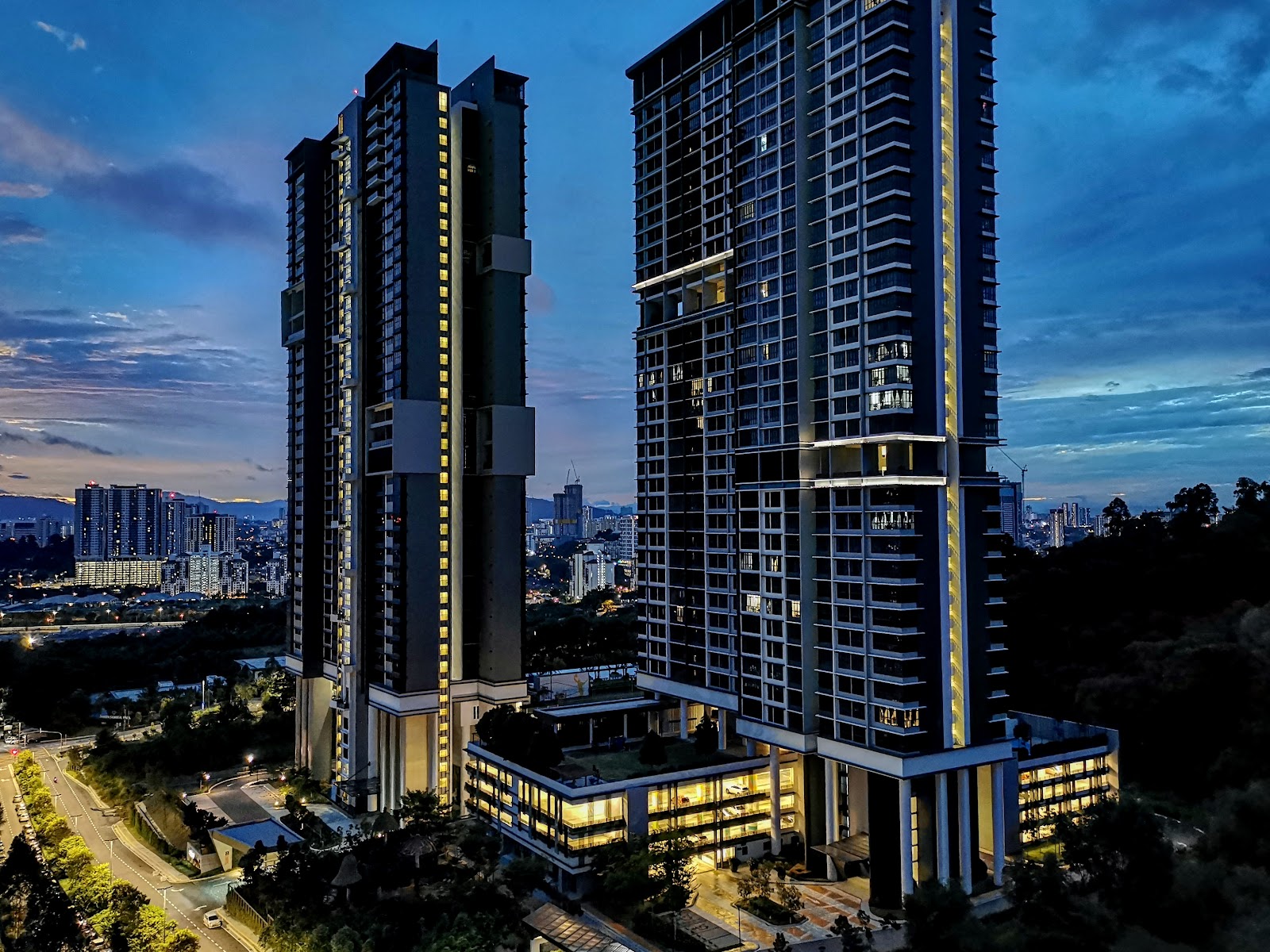 Inwood Residences: Twilight aerial view of two tall residential towers with illuminated facades, city skyline and hills in the background