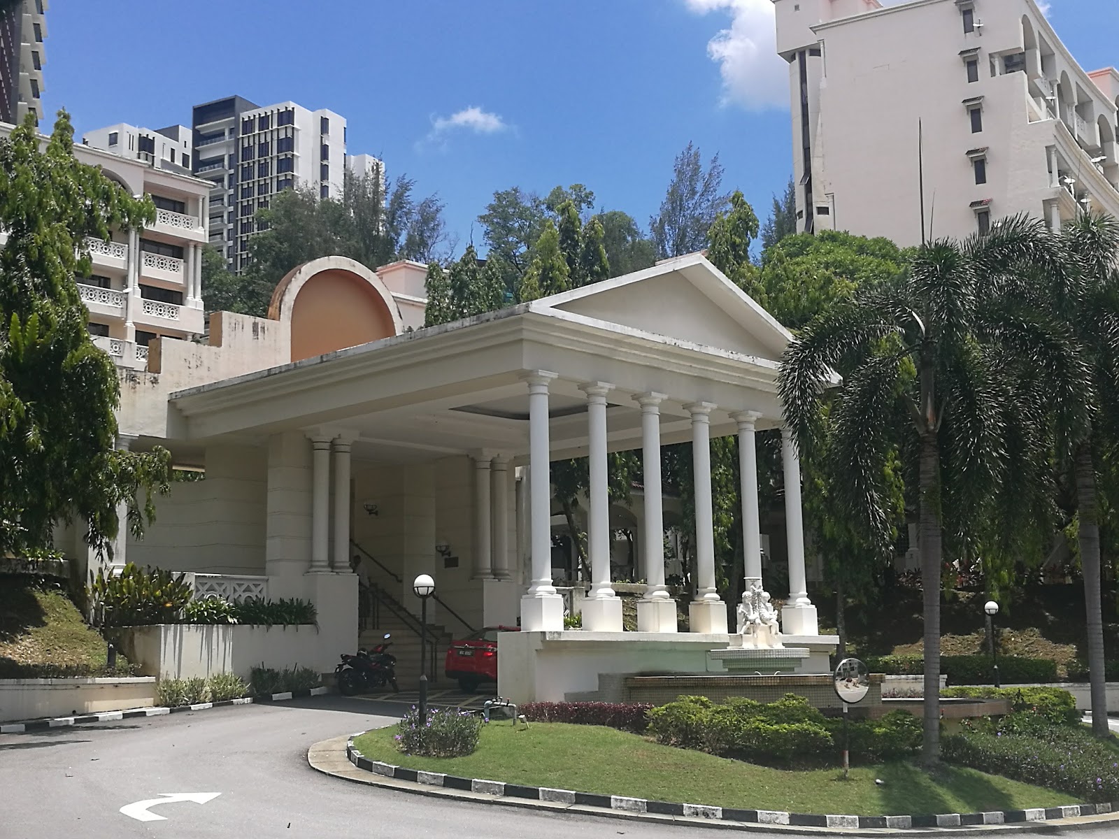 Tivoli Villas: Entrance porte-cochere with white columns and pediment roof at a residential development, surrounded by palm trees and other high-rise buildings, with a driveway and fountain