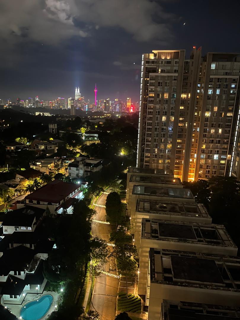 One Menerung: Night-time skyline view from a high-rise unit overlooking a neighborhood of houses, a swimming pool, and the distant illuminated city skyline including the Kuala Lumpur Tower and Petronas Towers