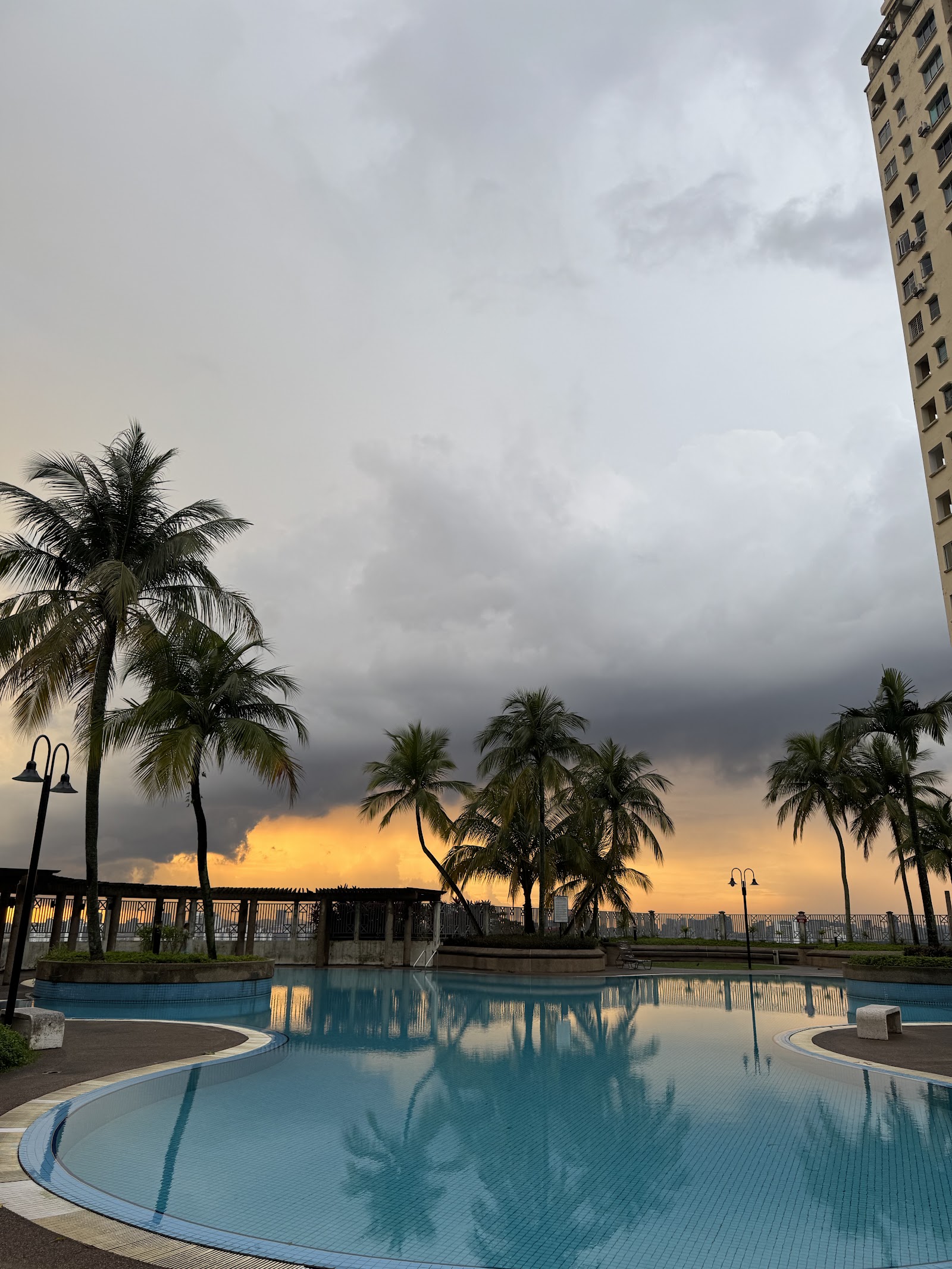 Pantai Panorama Condominiums: Infinity pool at sunset with palm trees and a dramatic cloudy sky, high-rise building visible on right