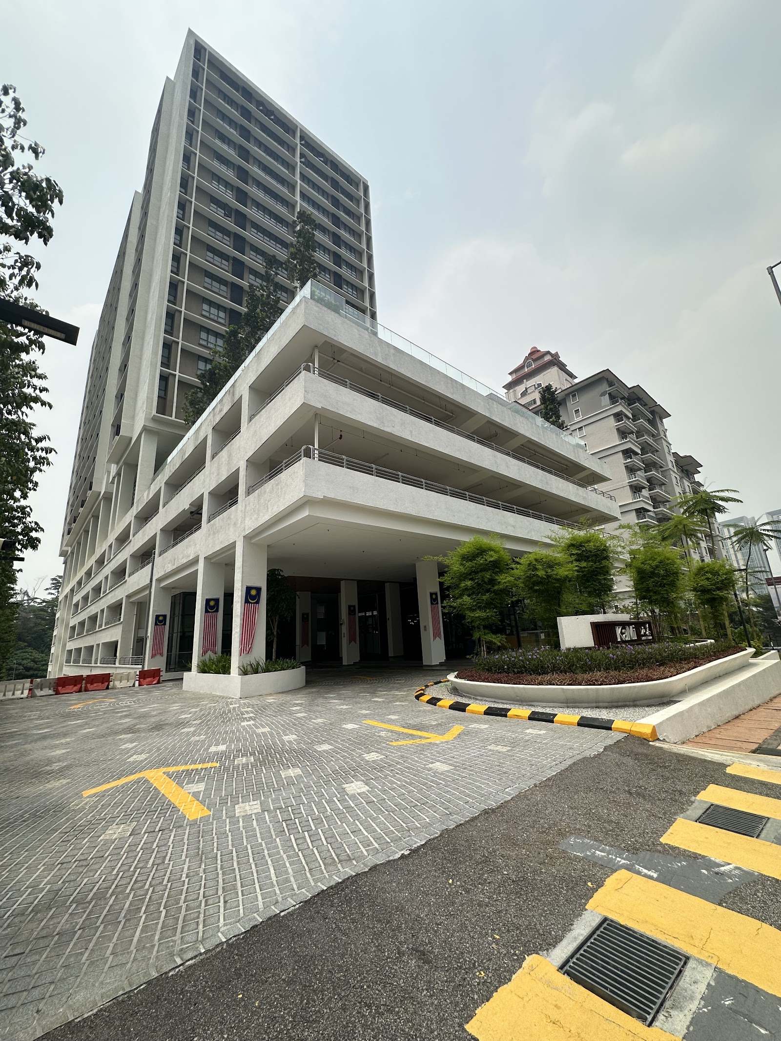 Kami: A high-rise residential building entrance with driveway, parking podium levels, and decorative Malaysian flags at the facade