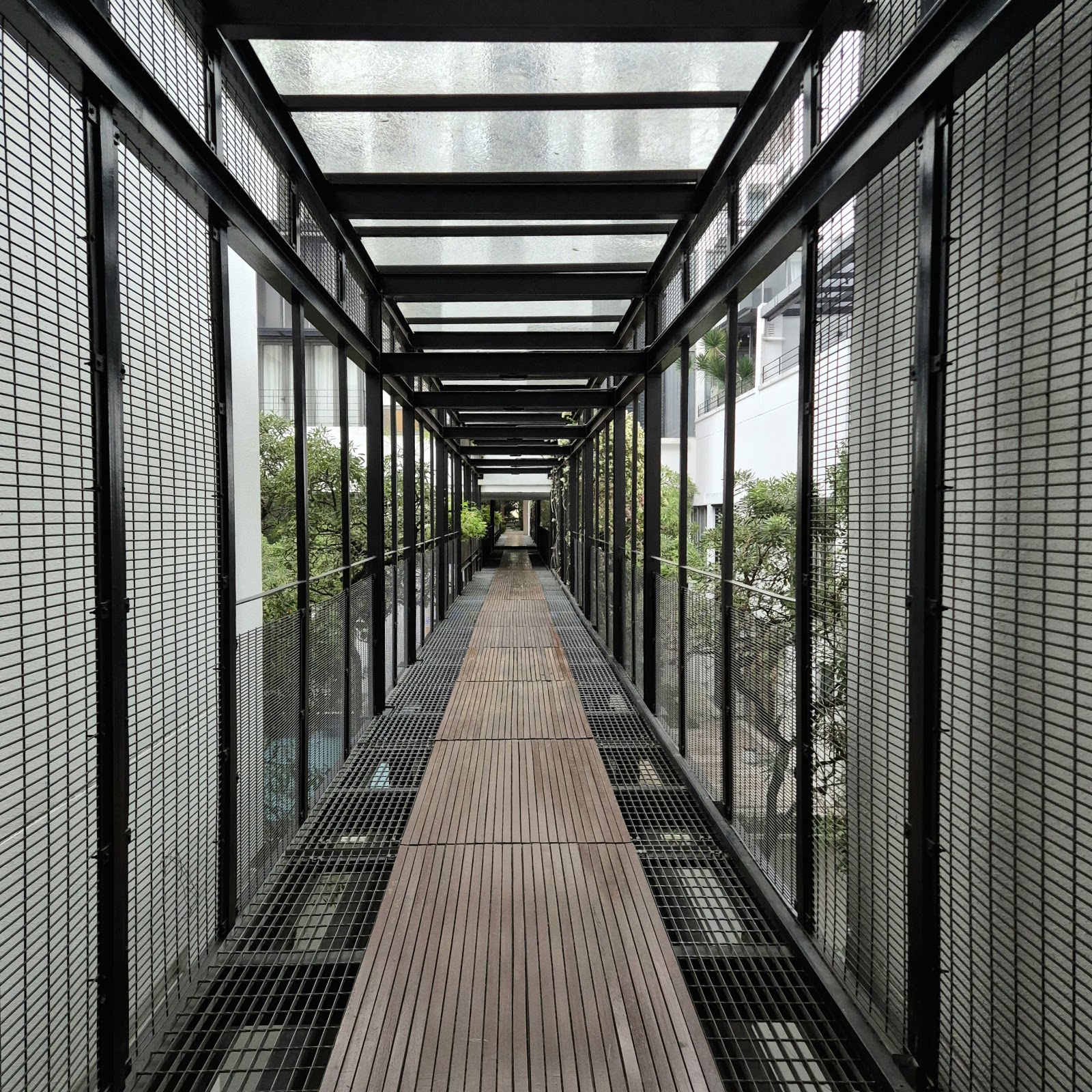 Centrio: A covered walkway with wooden decking, metal mesh side panels, and glass roof connecting parts of the development, with greenery visible through the mesh