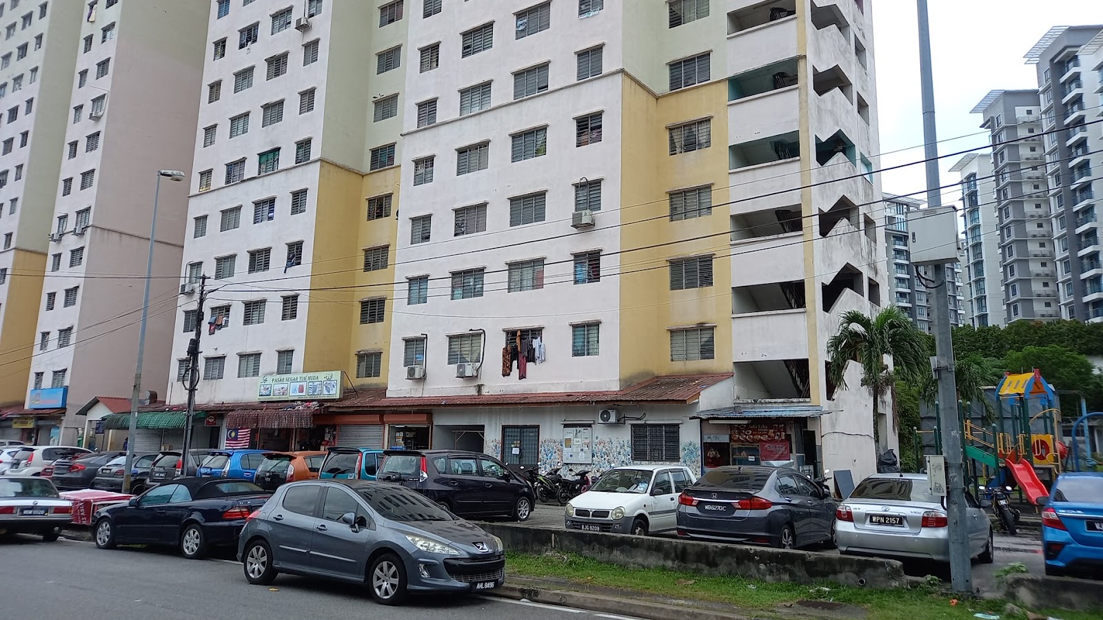 Sri Ara Apartment: Street view of a high-rise apartment building exterior with parked cars along the road, ground-floor shops, and a small playground nearby