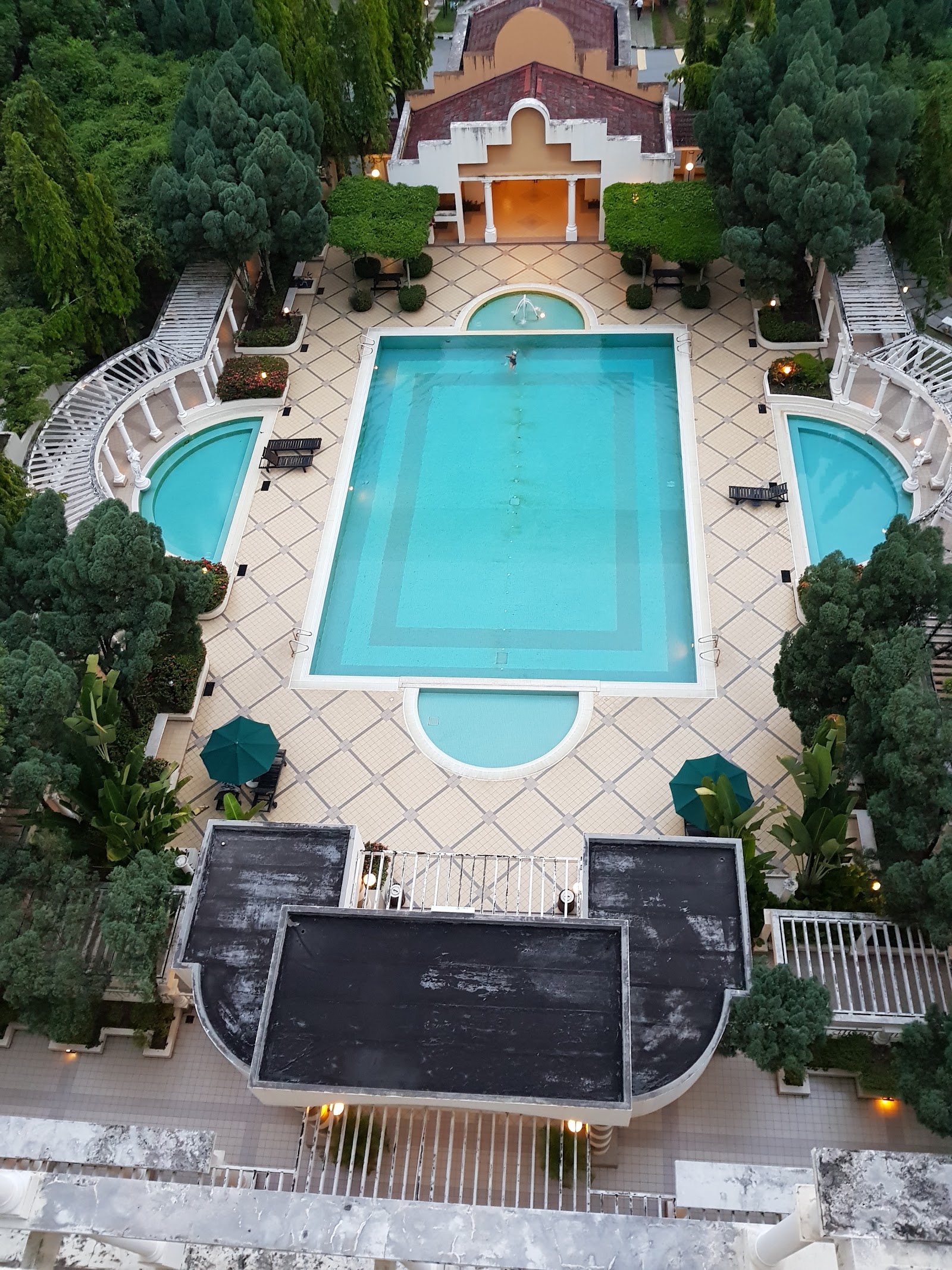 Tivoli Villas: Aerial view of an outdoor swimming pool complex with a rectangular main pool and two curved side pools, surrounded by tiled deck, umbrellas, and lush trees
