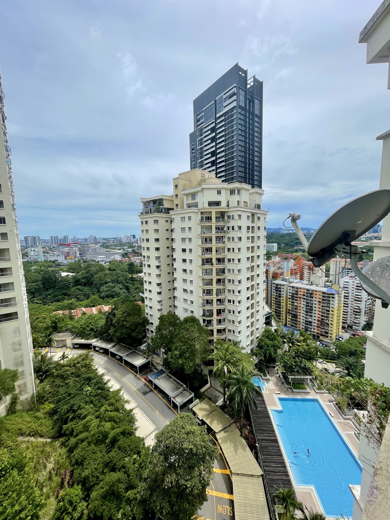Pantai Panorama Condominiums: Aerial view from a high-rise balcony showing a nearby residential tower, swimming pool below, dense trees, and city skyline in the background