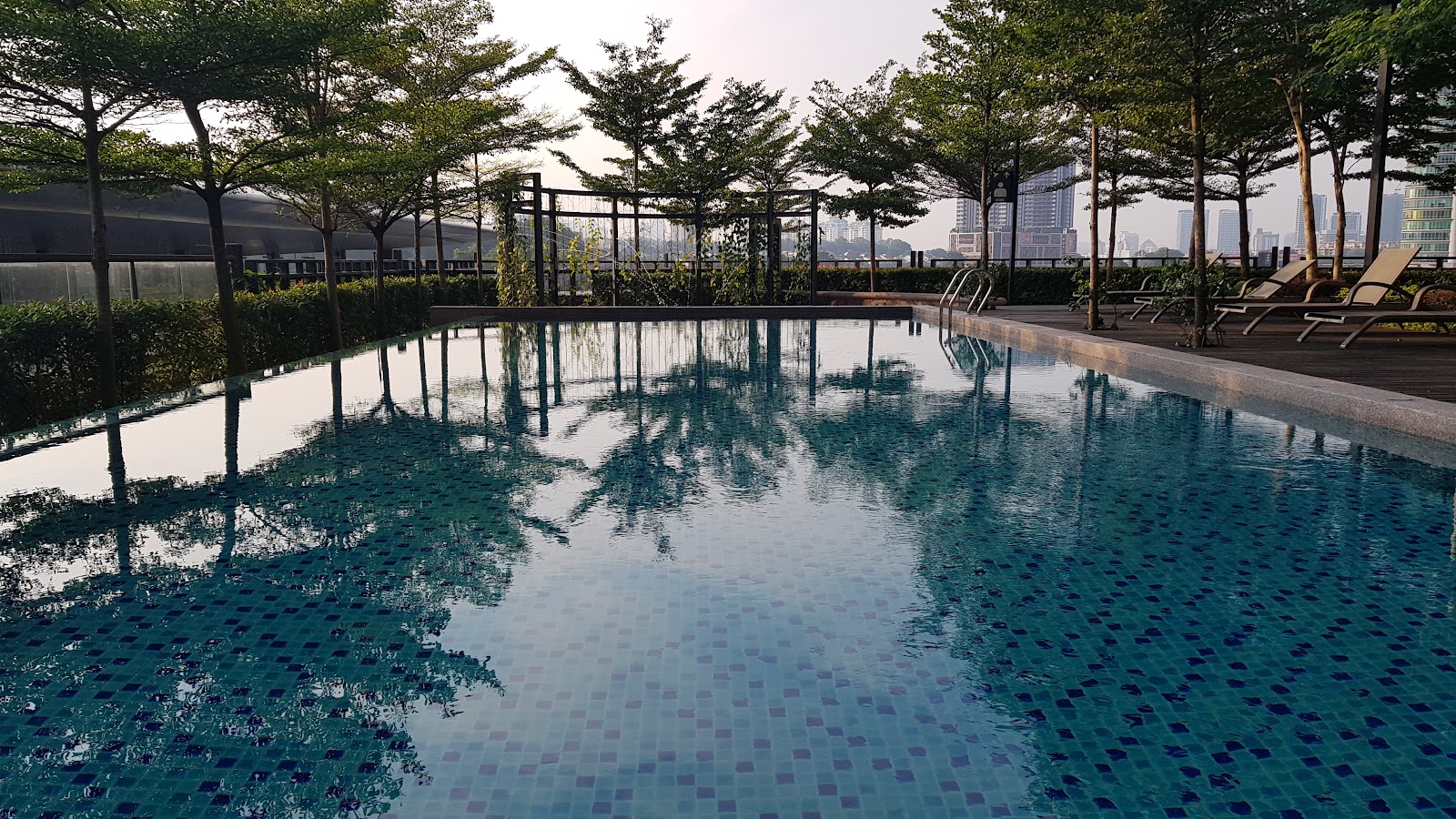 TTDI Ascencia: Rooftop infinity-edge swimming pool with tiled deck, sun loungers, surrounded by trees and a city skyline in the background