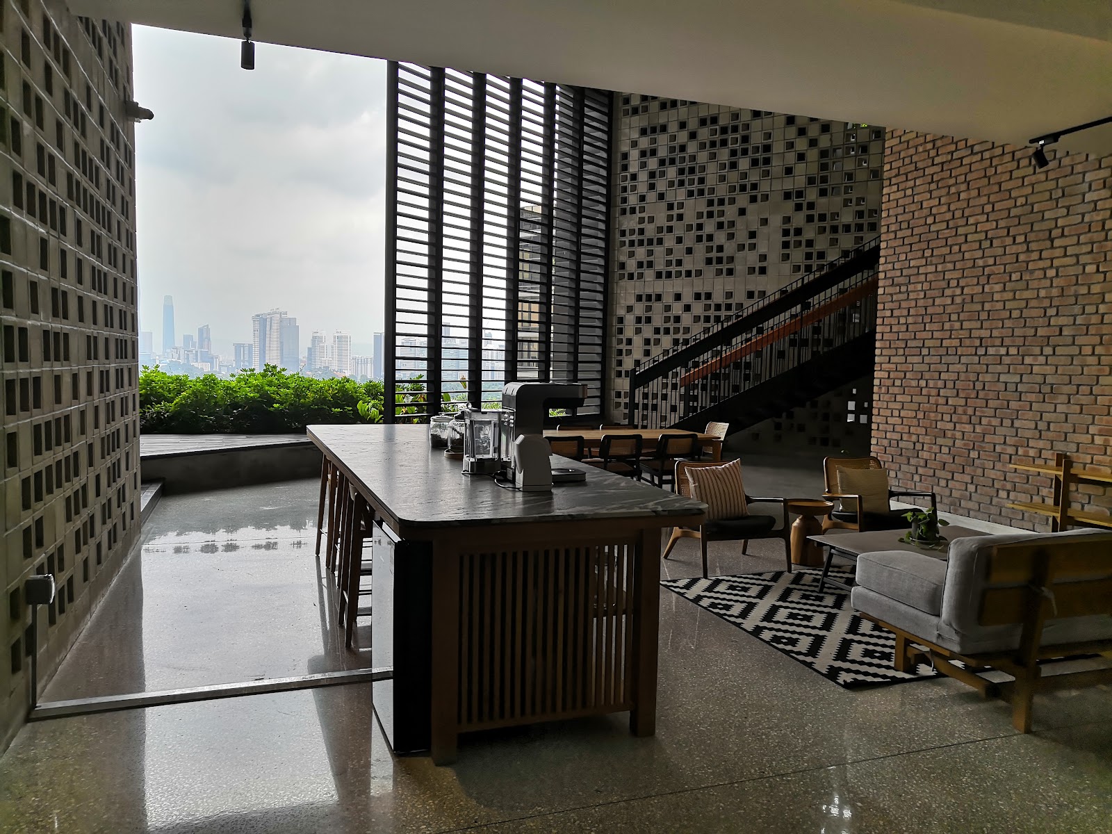 Kapas Heights: A lounge/lobby seating and bar area with a city skyline view through floor-to-ceiling windows, featuring a wooden table, sofas, and a staircase with patterned walls