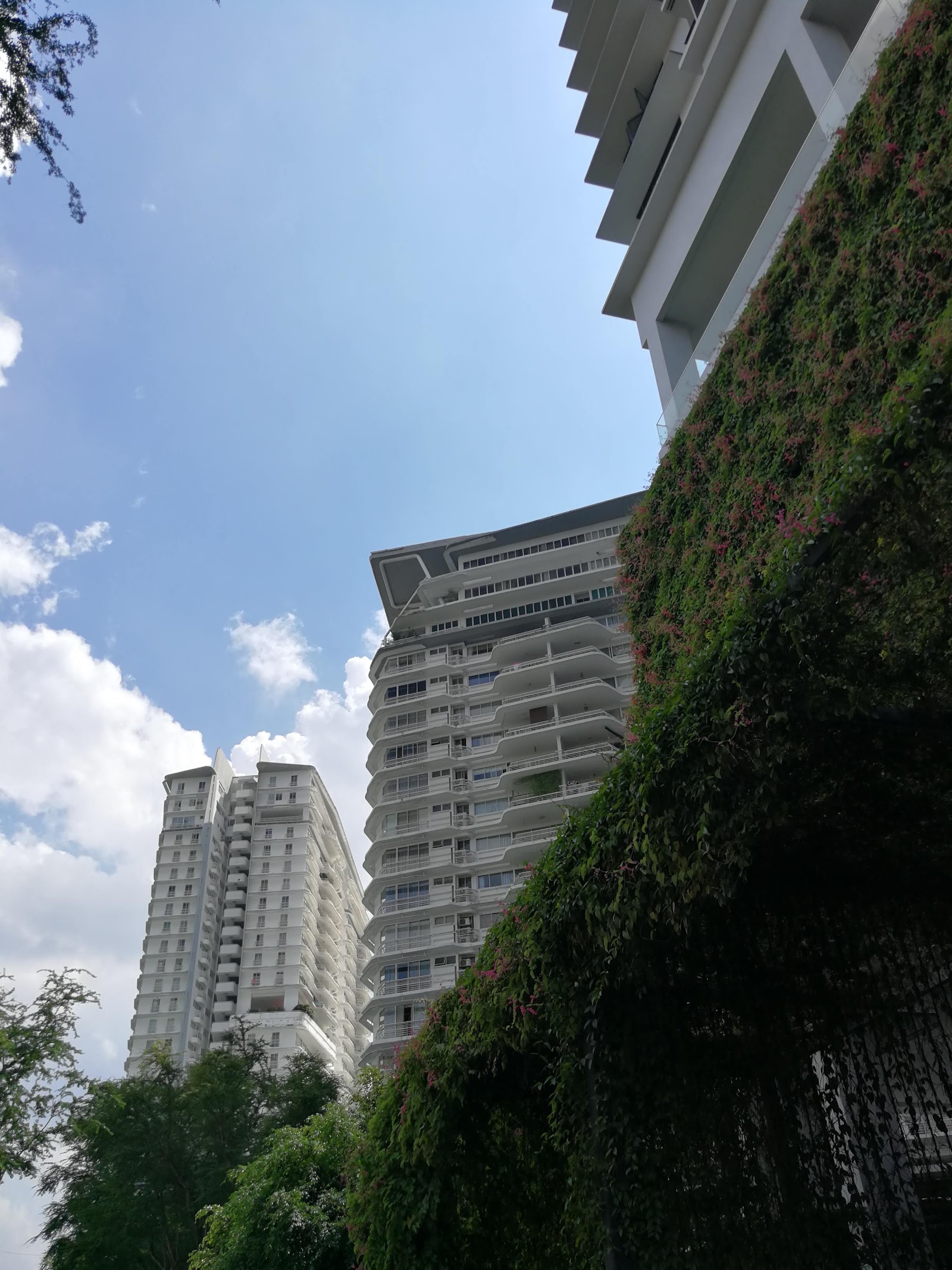 The Residence Condominium @ TTDI: Low-angle view of high-rise residential towers with balconies, seen past a wall covered in flowering vines and trees, under a partly cloudy sky