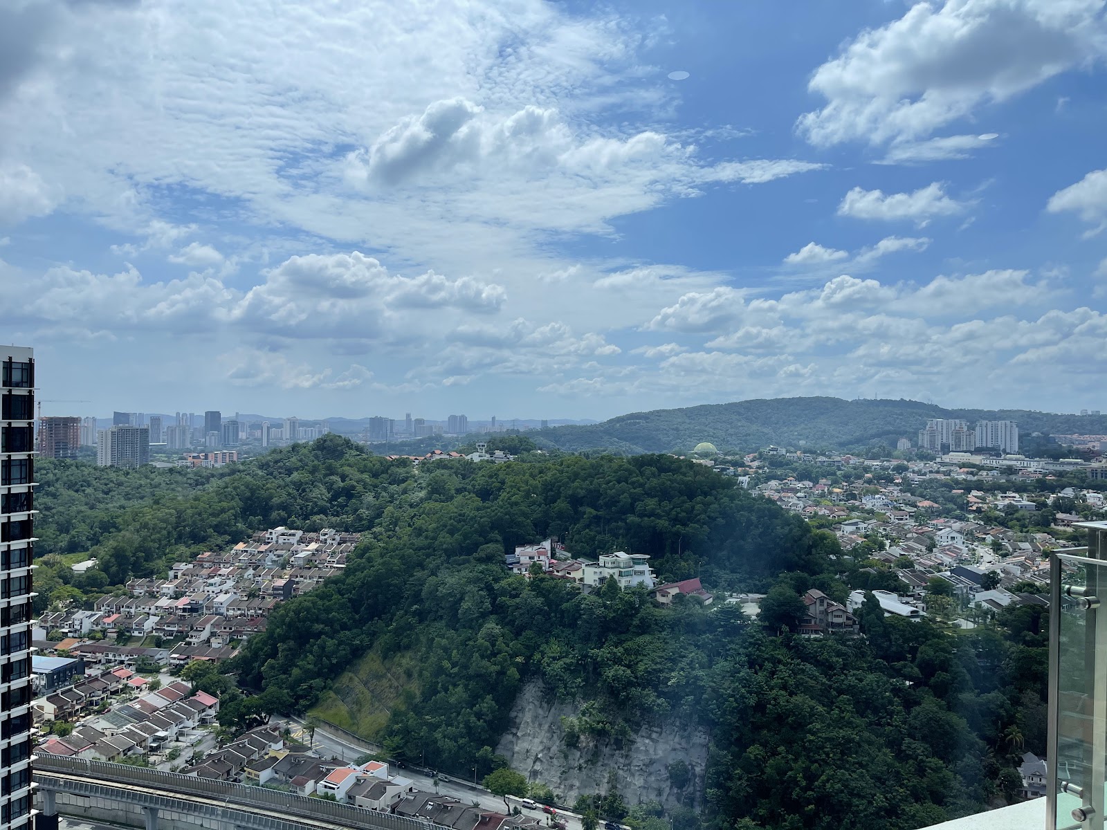 Serai: Panoramic view from a high-rise balcony overlooking a hillside neighborhood with trees, houses, distant city skyline and hills under a partly cloudy sky