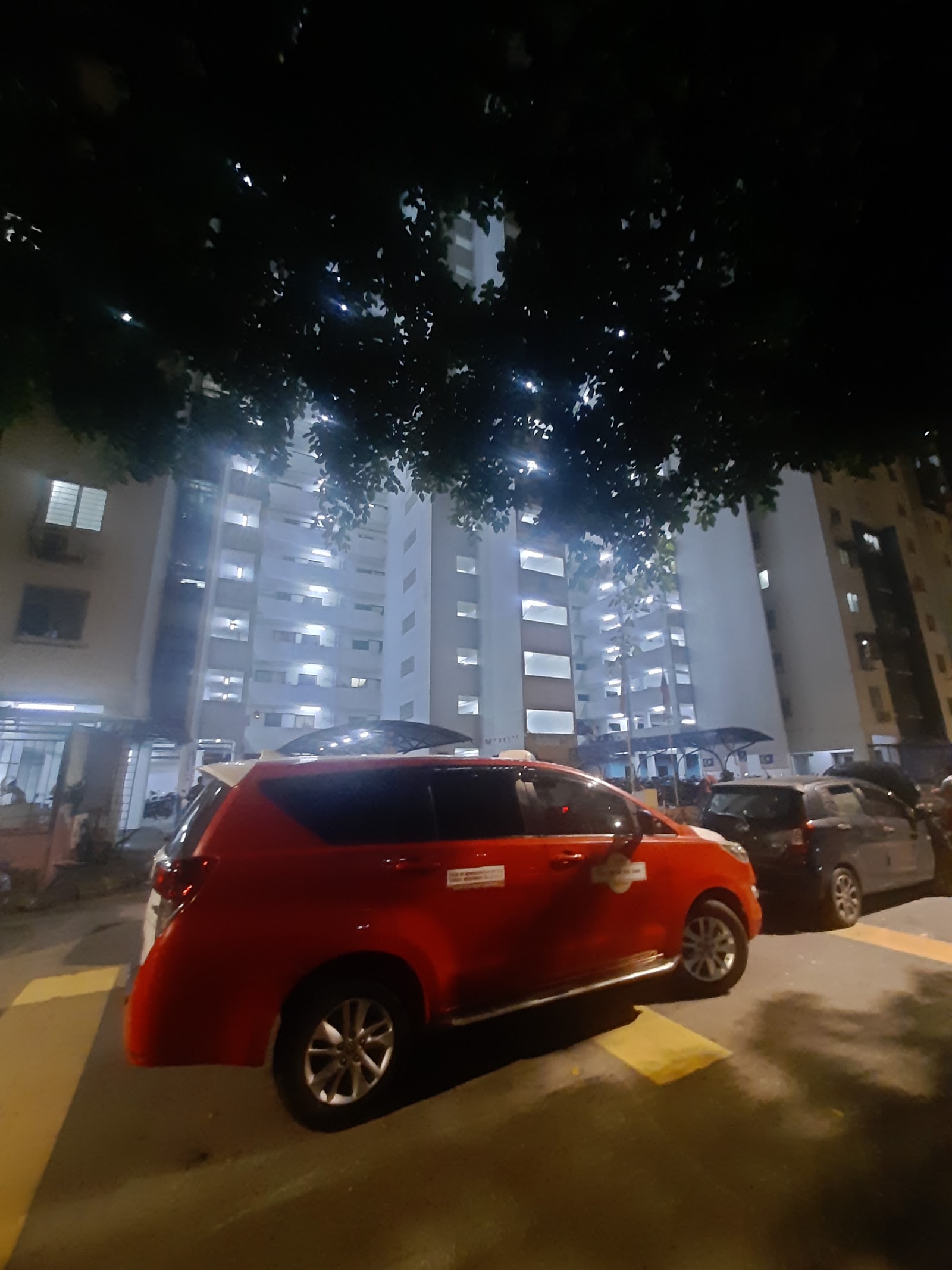 PPR Pantai Ria: Night view of apartment towers with a red car parked in front, seen through tree branches