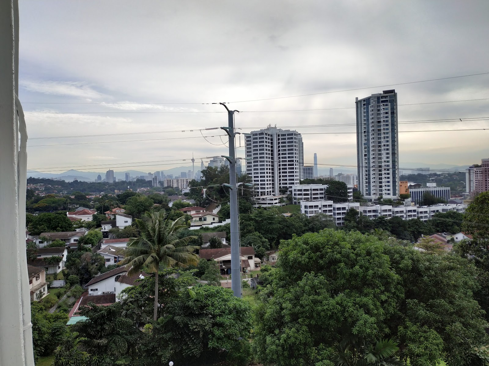 Pantai Towers: View from a high floor overlooking treetops, low-rise houses, and city skyscrapers under an overcast sky