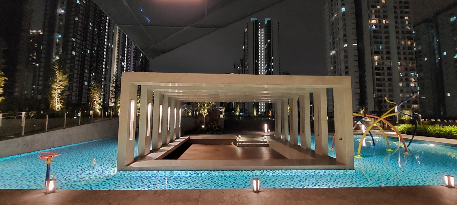 Trinity Pentamont: Nighttime view of a rooftop swimming pool with a covered pergola walkway, surrounded by illuminated high-rise buildings