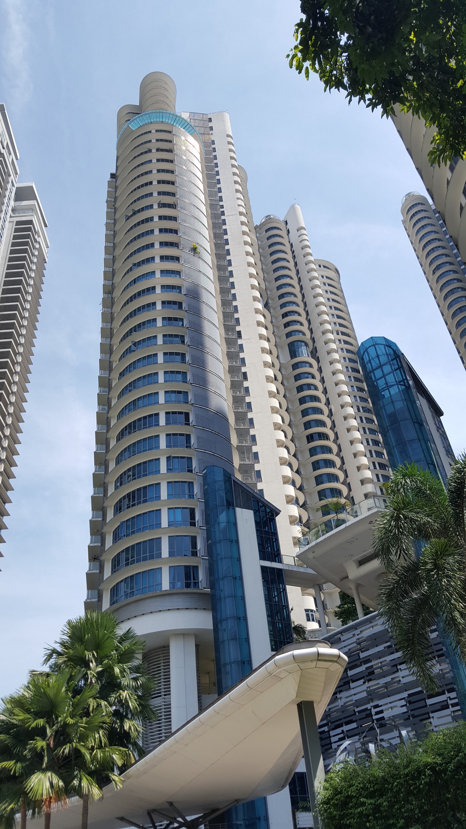 Mont Kiara Pelangi: Upward view of tall curved residential towers with distinctive balcony railings, seen from ground level among palm trees
