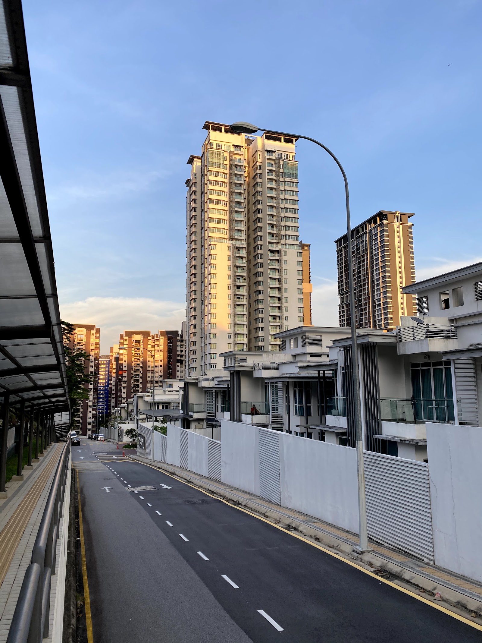 Saville @ The Park Bangsar: A street view showing high-rise residential towers and townhouses along a road with a covered walkway in the foreground, taken at sunset