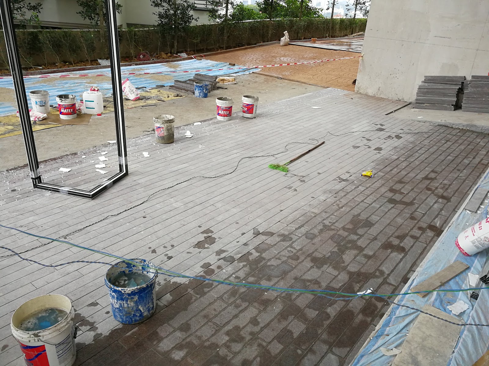 Impian Emas: Construction site under a covered walkway with paint buckets, a broom, tools, and building materials scattered on the tiled floor
