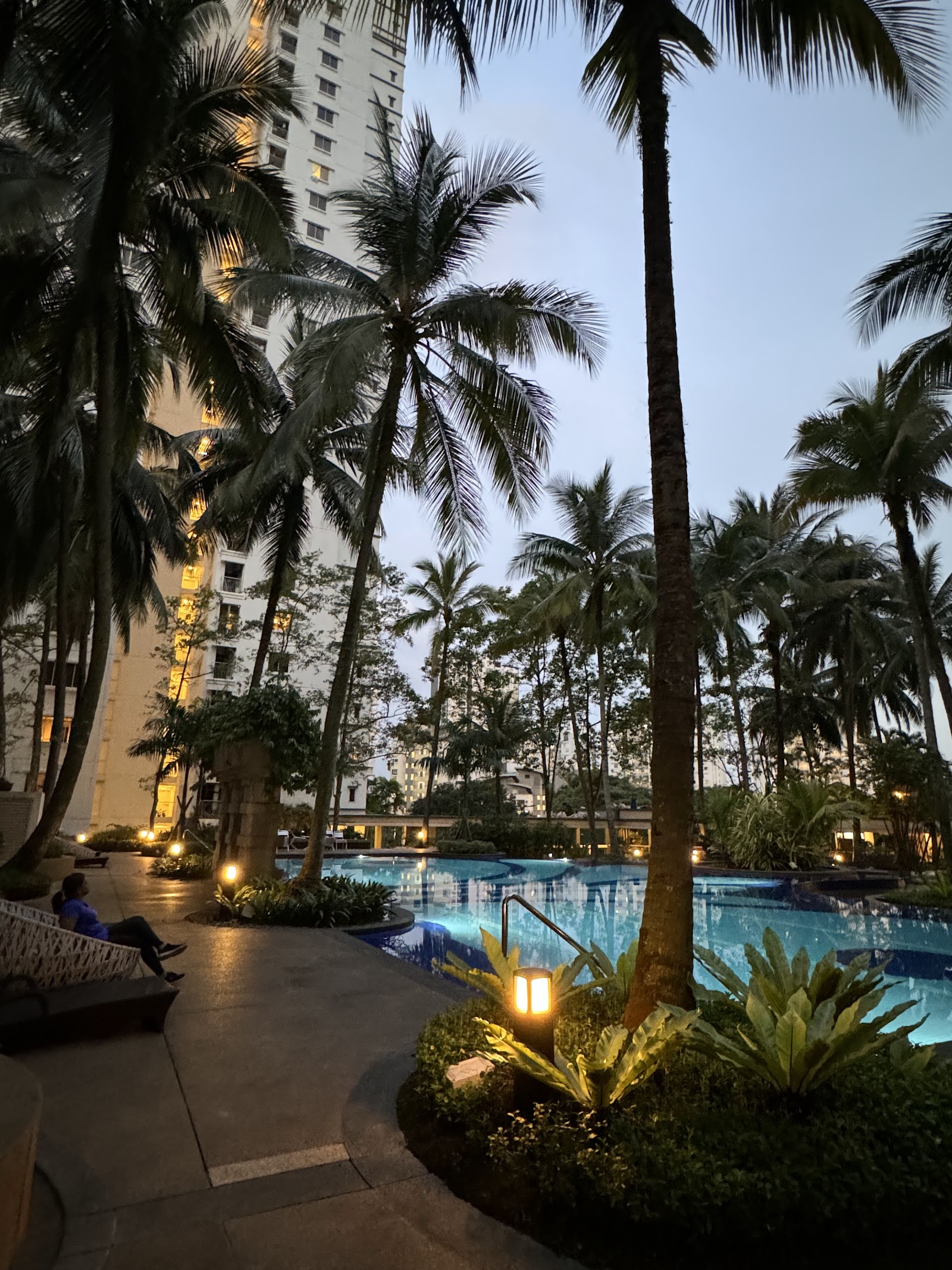 Kiaramas Sutera: Outdoor swimming pool area surrounded by tall palm trees with residential towers in the background at dusk