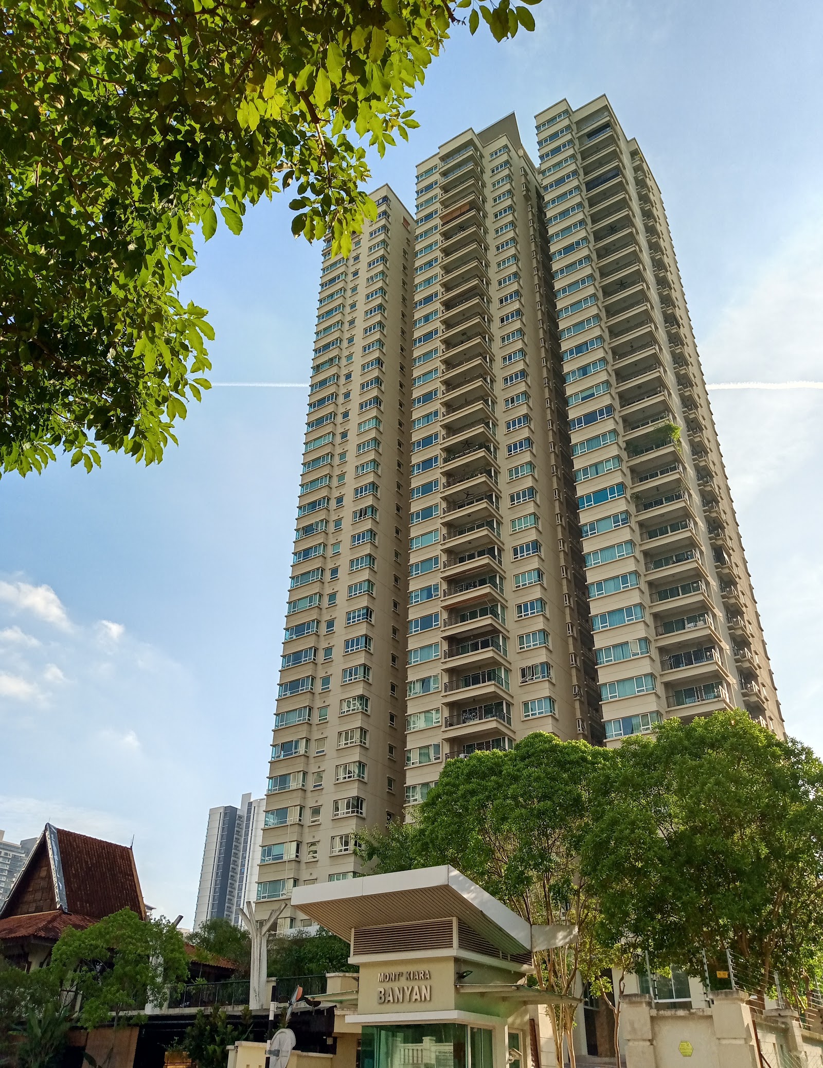 Mont Kiara Banyan: A tall residential tower of Mont Kiara Banyan viewed from ground level with trees in foreground and a signage kiosk at the base