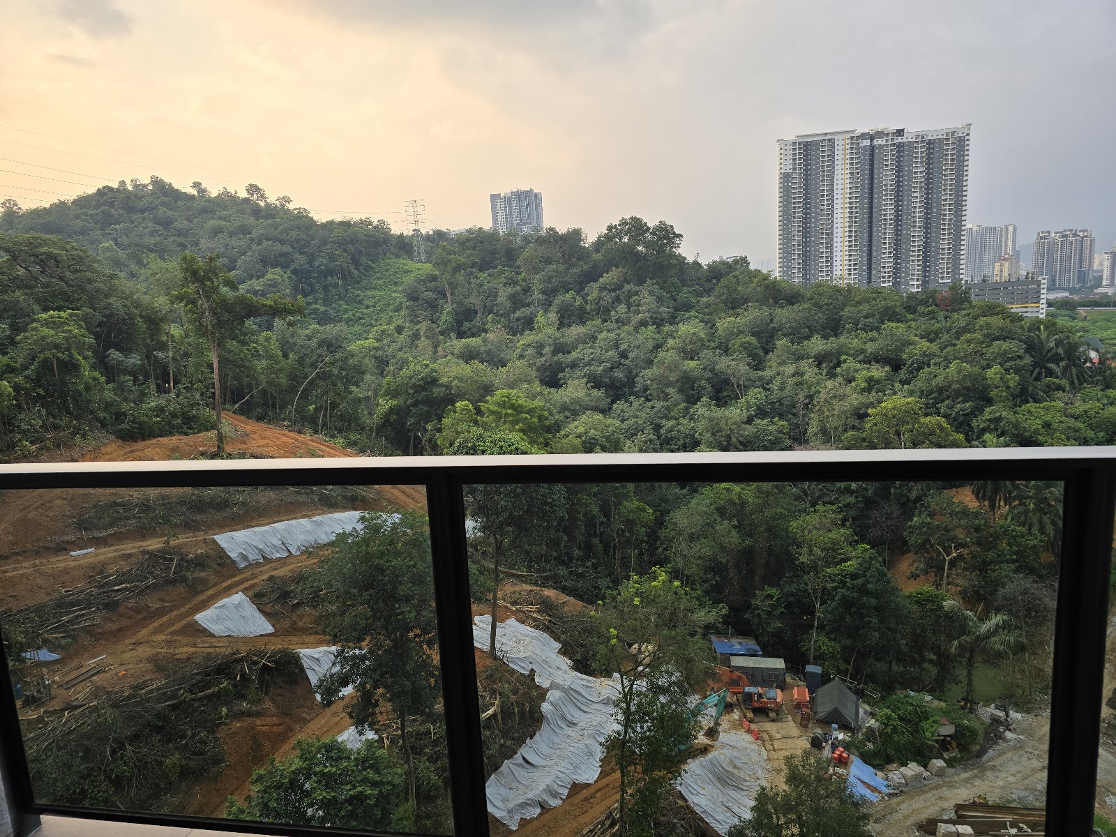 Residensi Astrea: View from a high-rise balcony overlooking dense forest, a construction site with tarps, and distant city high-rise buildings