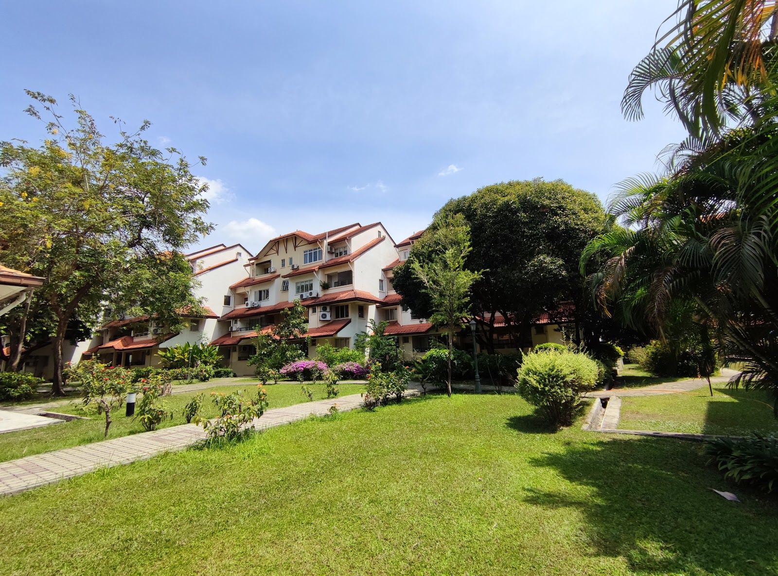 Villa Flora: Exterior view of a low-rise residential apartment complex with red-tiled roofs, surrounded by landscaped gardens, trees, and shrubs under a blue sky