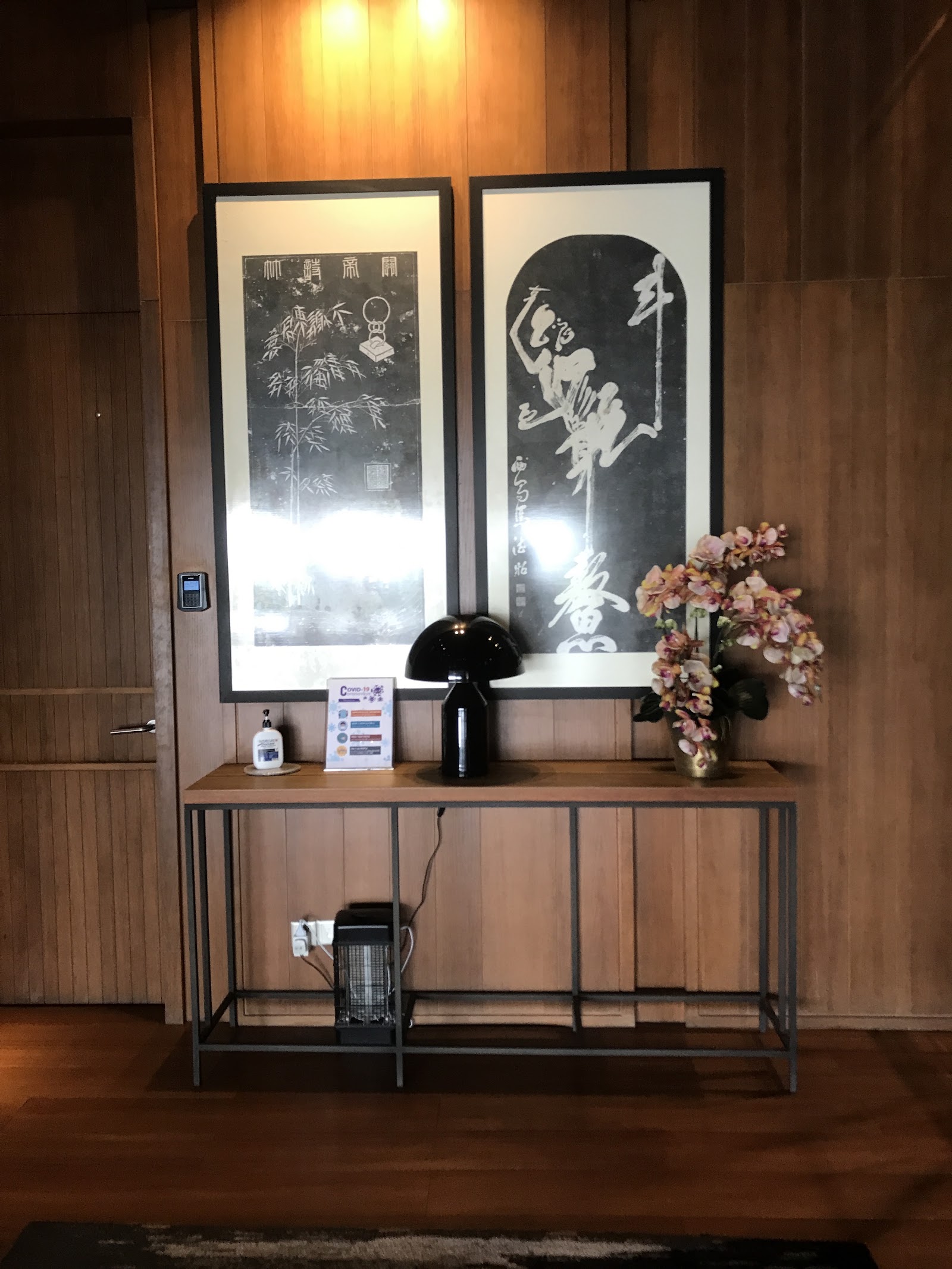 Rhombus @ Bangsar: A lobby console table with framed calligraphy artwork, a lamp, flowers, and a sanitizer station against wood-paneled walls