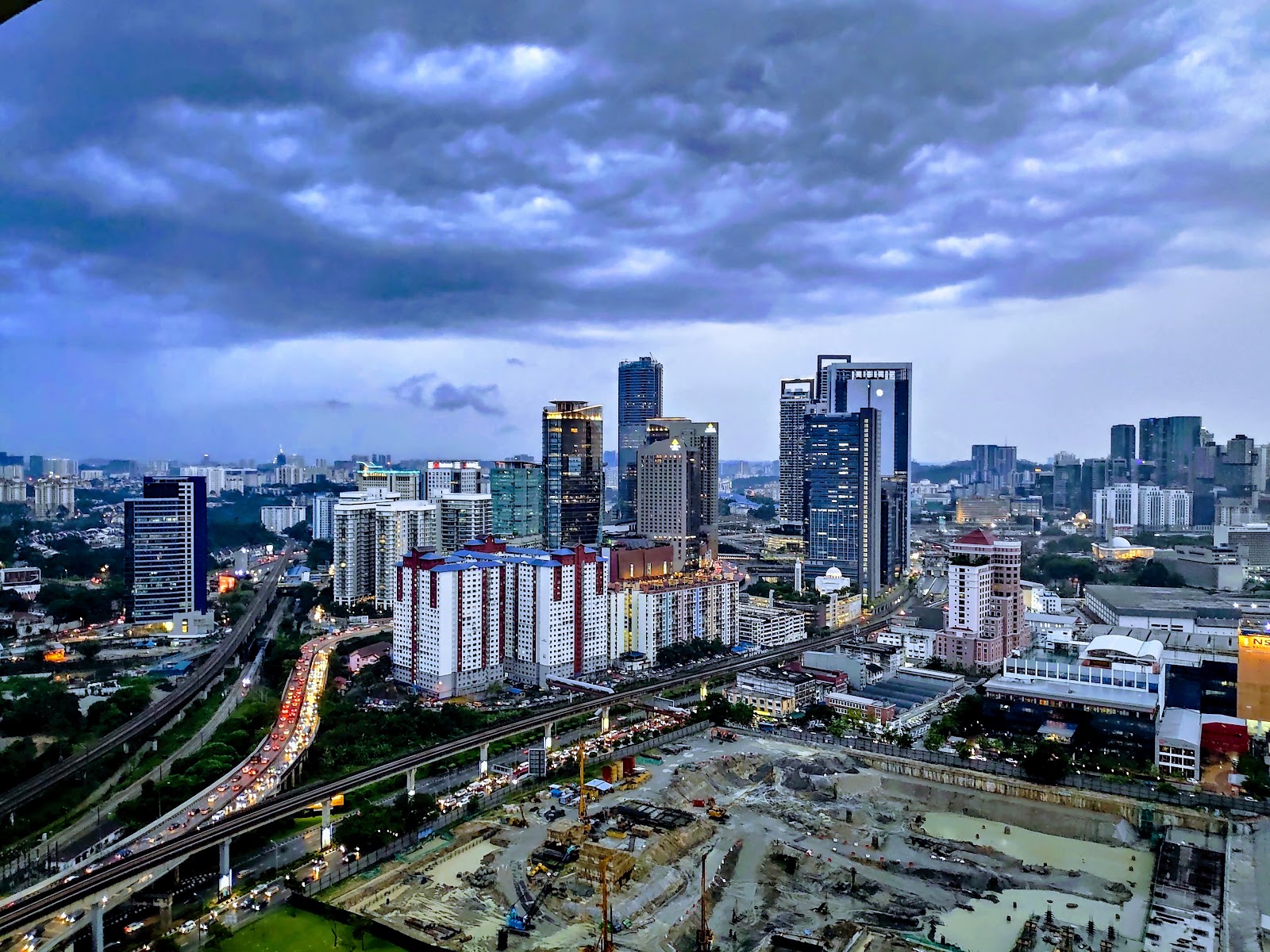 Gaya Bangsar: Aerial dusk view of a city skyline with skyscrapers, construction site, and a highway from a high vantage point