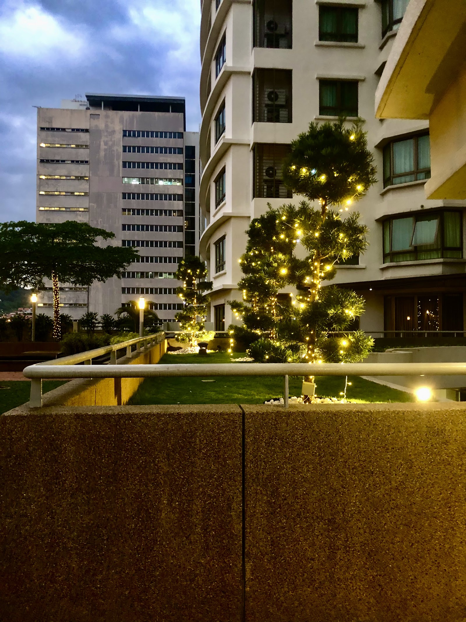 i-Zen @ Kiara 2: Evening view of a residential tower's garden courtyard with lit decorative trees, a lawn, and surrounding high-rise buildings in the background
