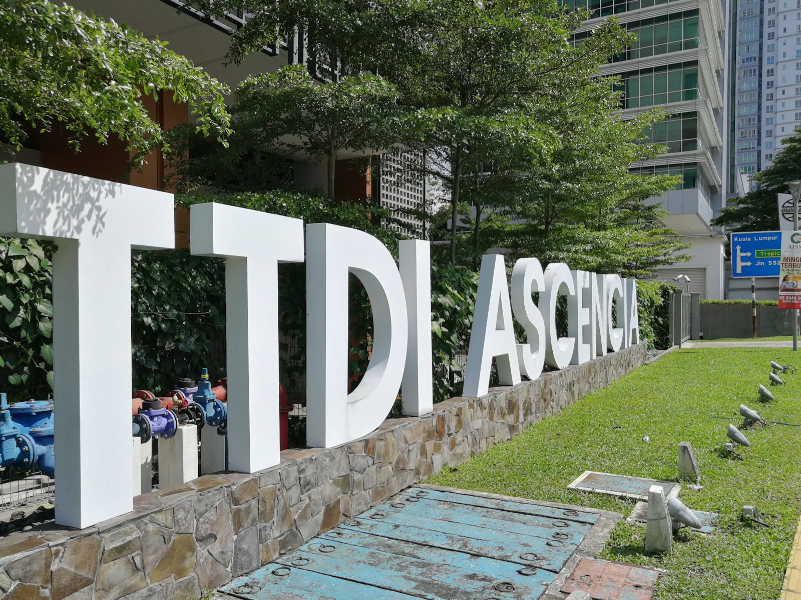 TTDI Ascencia: Large white 3D letters spelling 'TTDI ASCENCIA' installed on a stone wall at the entrance grounds of the development, with trees and city towers in the background