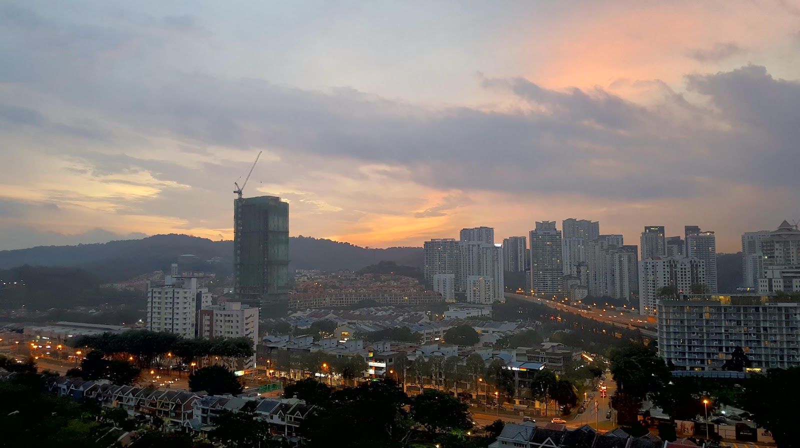 Menara Hartamas: City skyline at sunset viewed from a high vantage point, showing skyscrapers, a construction crane, residential buildings, and roads with lights