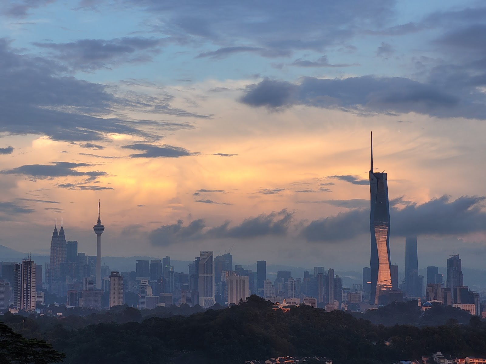 The Loft: City skyline at sunset with skyscrapers including KL Tower and Menara telecommunications tower silhouetted against colorful clouds
