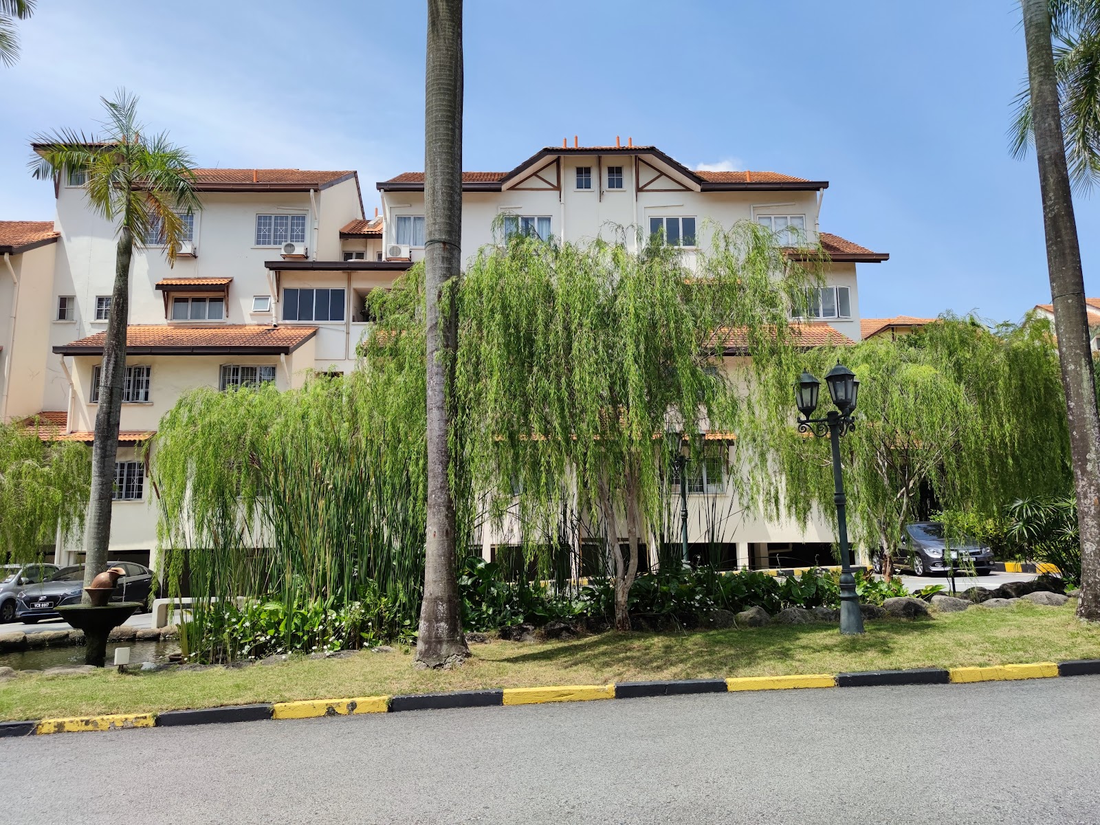 Villa Flora: Exterior view of a residential building with terracotta roofing, surrounded by weeping willow trees, palm trees, and a landscaped garden with a small fountain, parked cars visible near the building