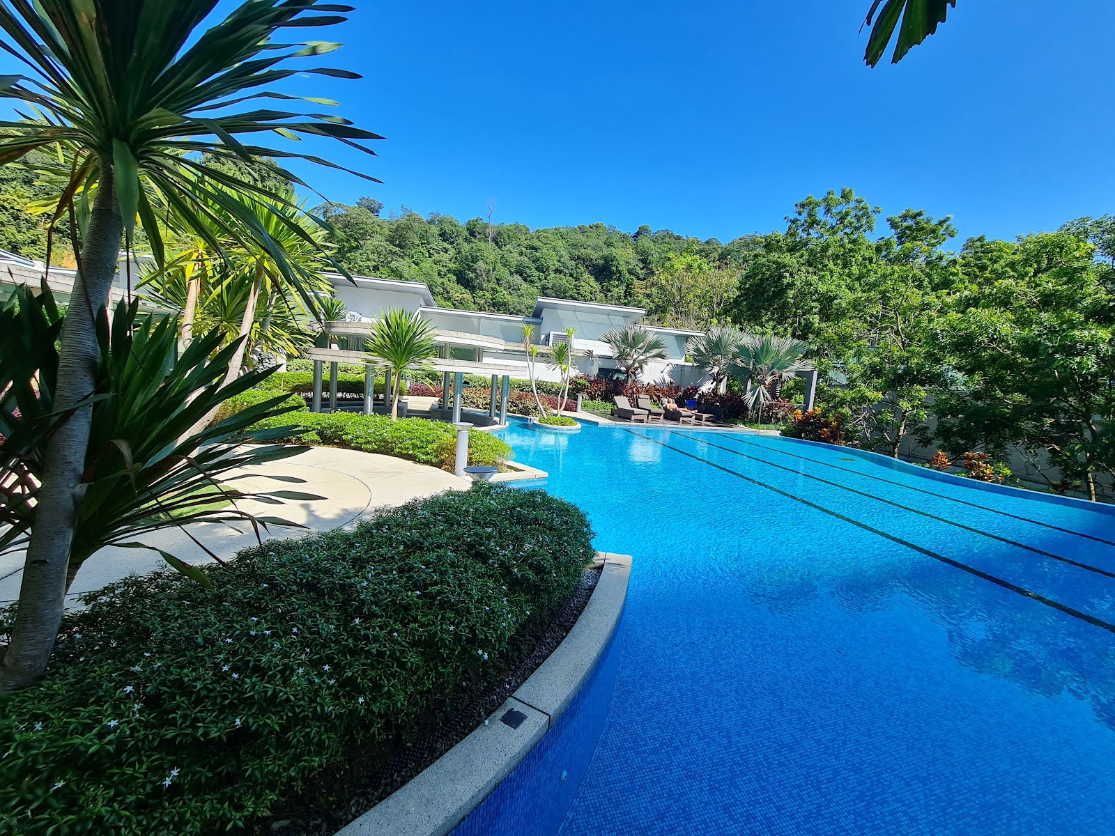 Kiaramas Cendana: An infinity-edge swimming pool surrounded by tropical landscaping, with low-rise residential buildings and forested hills in the background
