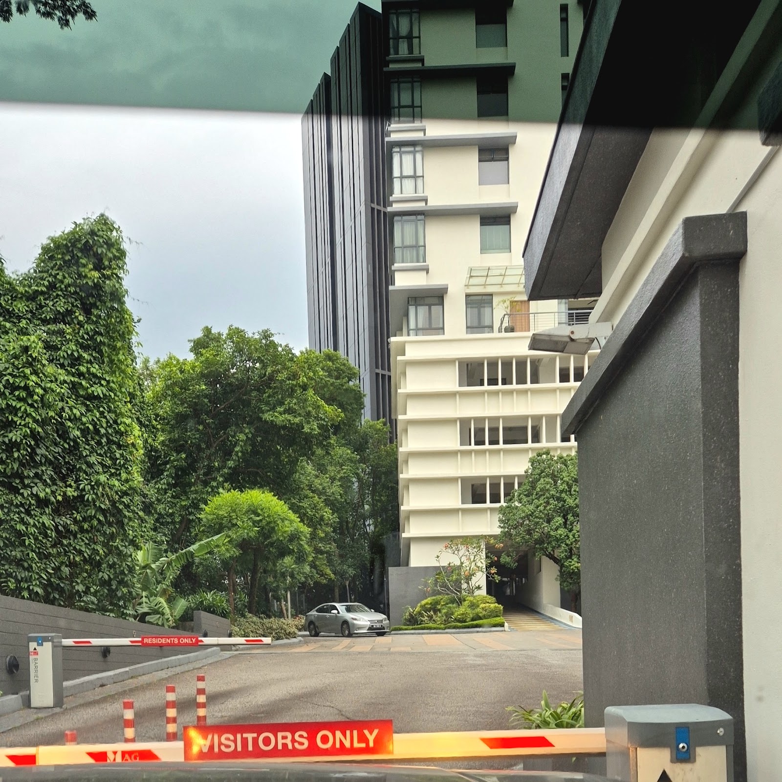 Bangsar Peak: View through a boom gate at a residential building entrance with barrier arms marked 'RESIDENTS ONLY' and 'VISITORS ONLY', driveway and greenery in foreground