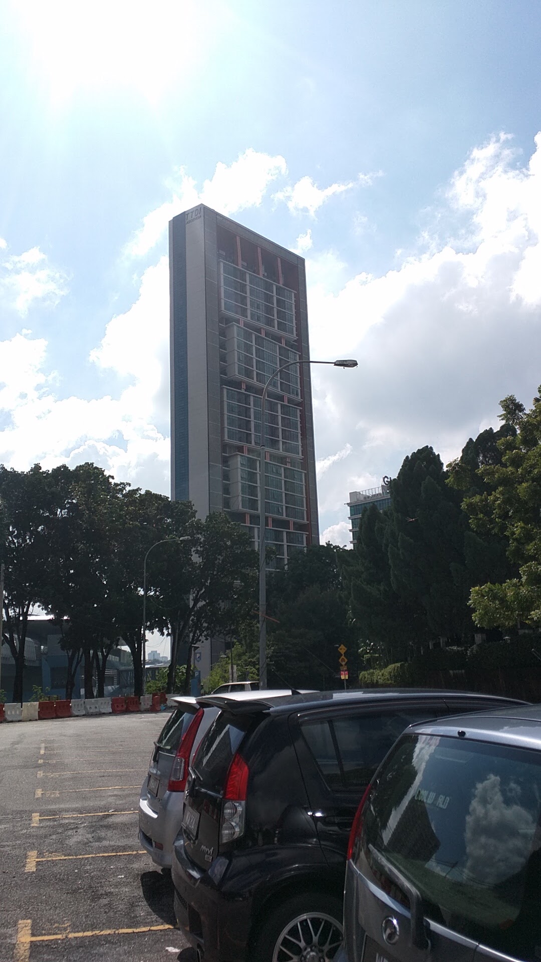 TTDI Ascencia: A tall high-rise residential building viewed from a street-level parking area with trees and parked cars in foreground