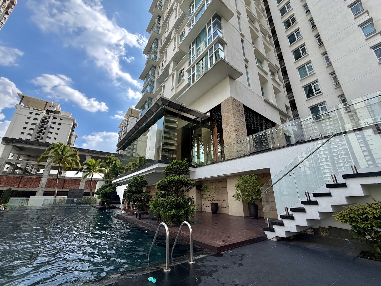 Kiara 9 Residency: Swimming pool with wooden deck and staircase at the base of a high-rise residential tower, with palm trees and neighboring buildings visible